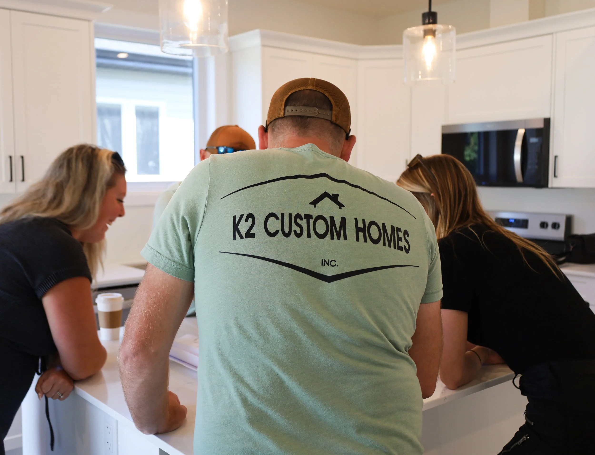 Four people gathered around a kitchen counter, examining documents or drawings, with kitchen appliances visible in the background.