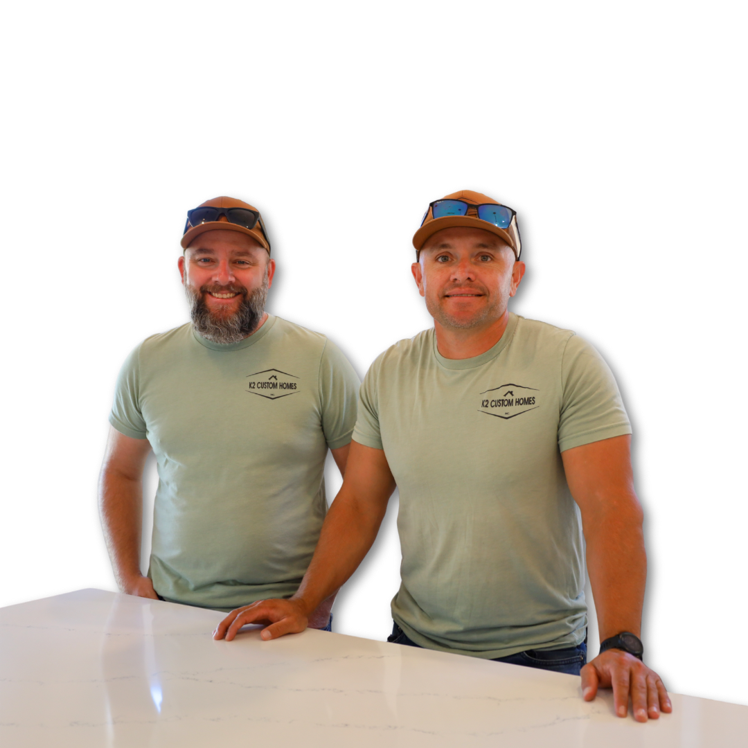 Two men in light green t-shirts and baseball caps with sunglasses on top, standing behind a white table, smiling at the camera, with a black background.