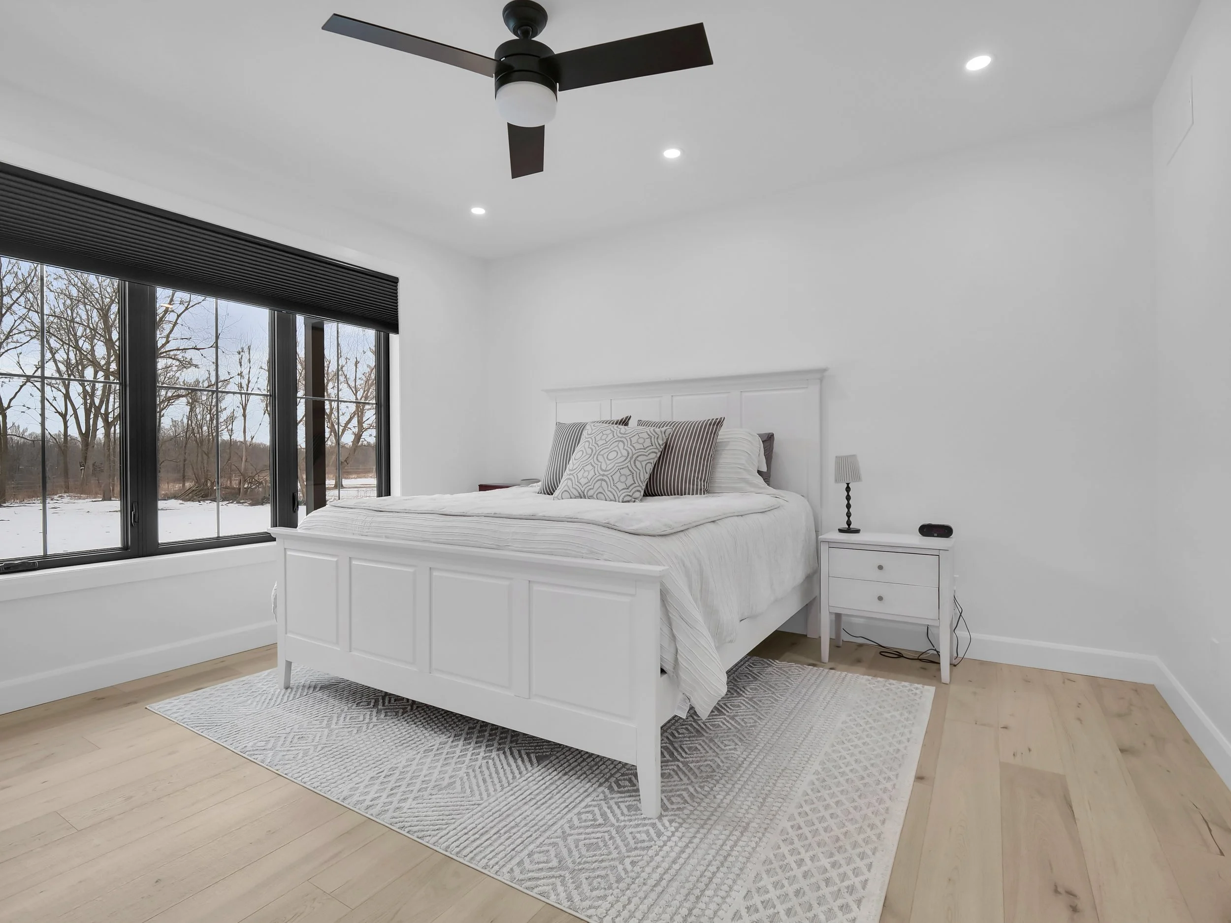 A minimalist white bedroom with a large bed, black window blinds, a ceiling fan, small bedside table with a lamp, and a view of snow outside.