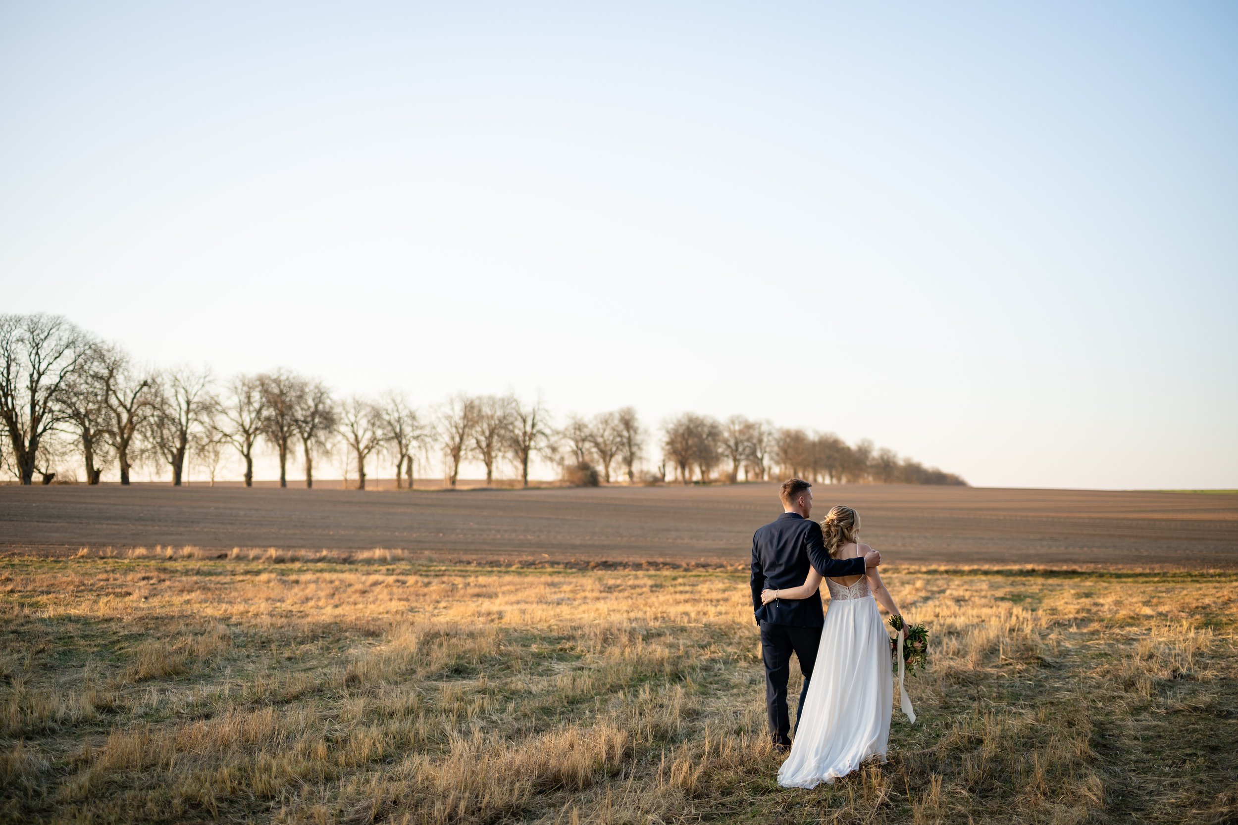 Ein Brautpaar spaziert während eines Sonnenuntergangs auf einem Feld in Brandenburg in der Nähe von Berlin in Hochzeitskleid und Hochzeitsanzug. Aufgenommen von Hochzeitsfotograf Dominic Glock.