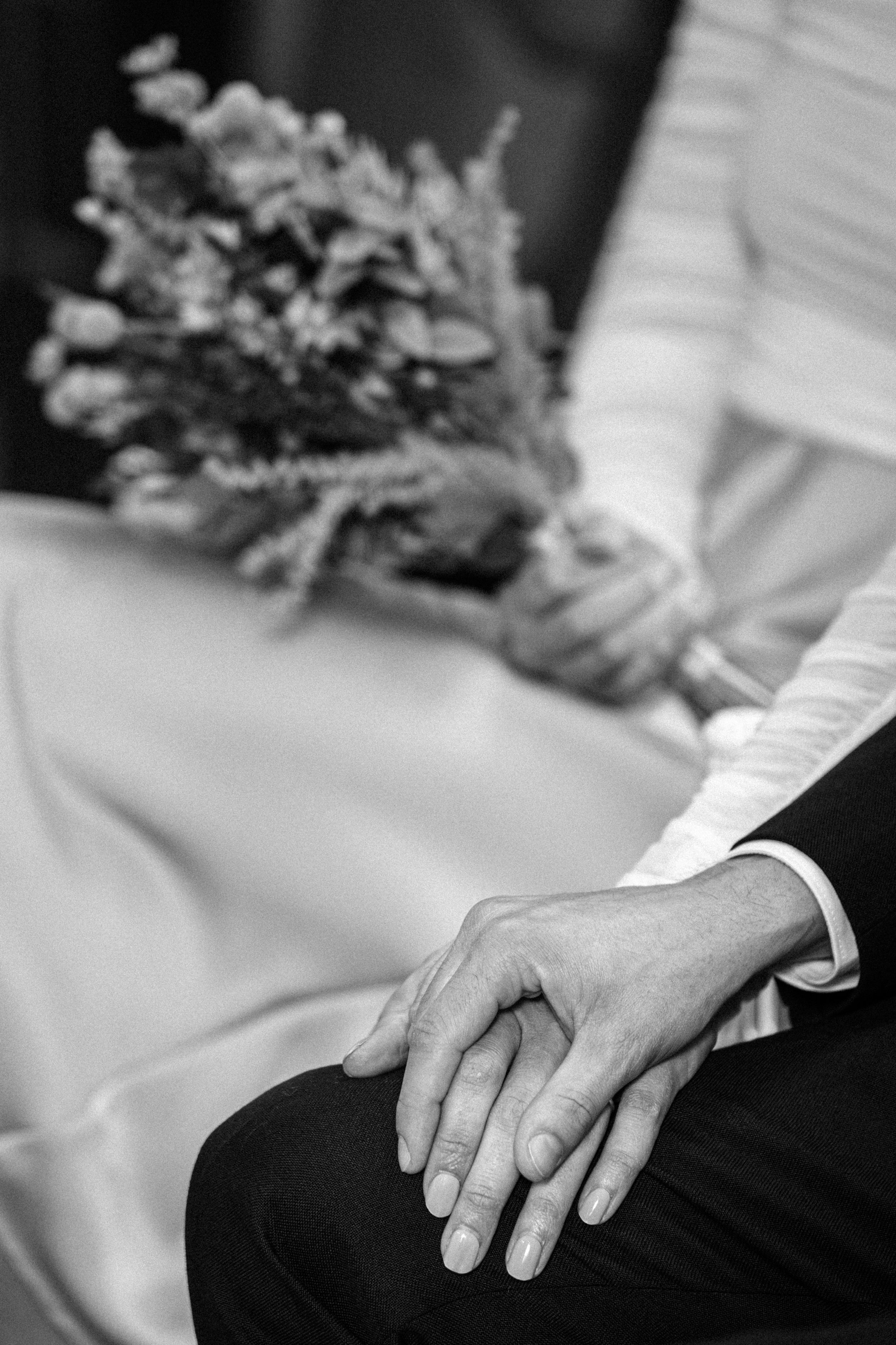 Nahaufnahme von zwei sich haltenden Händen bei einer Hochzeitsfeier, im Hintergrund ein Brautstrauß.