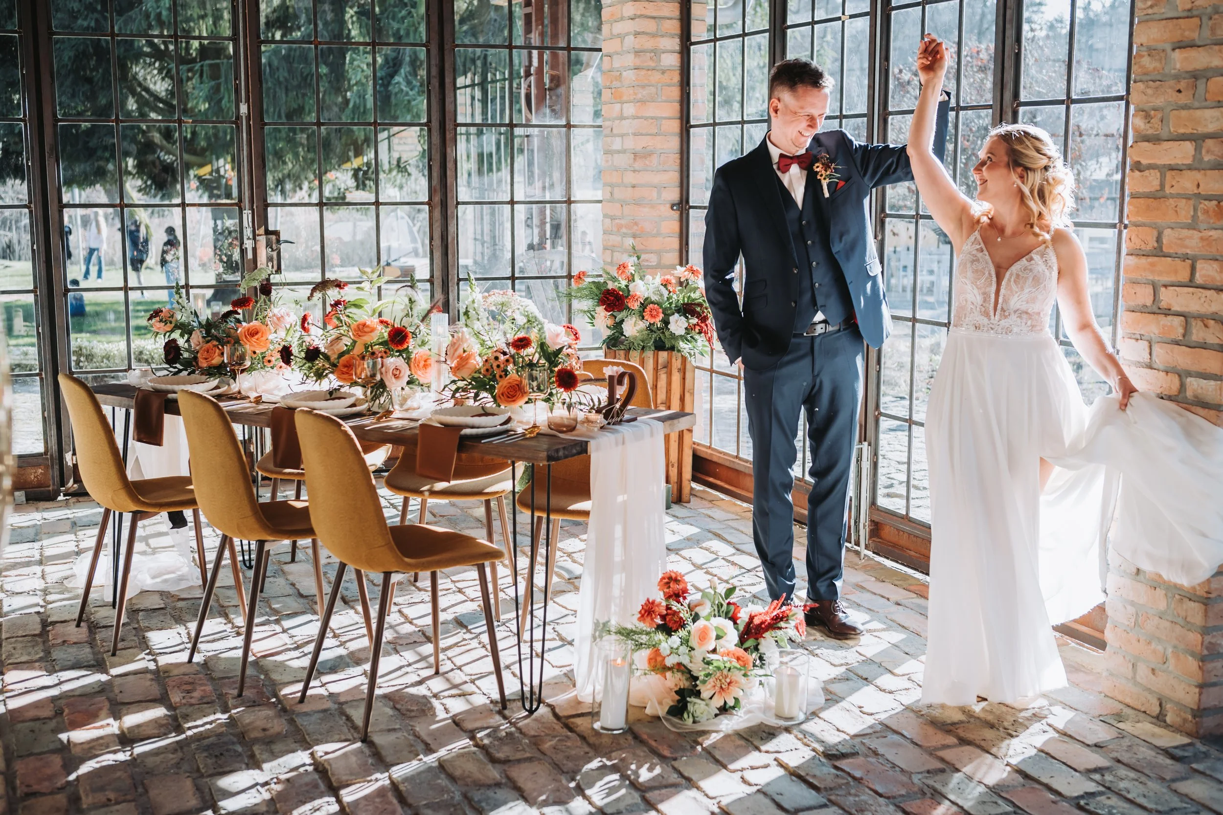 Ein Brautpaar tanzt bei einer Hochzeit in einem Raum mit großen Fensterflächen. Der Tisch ist festlich mit Blumen dekoriert.