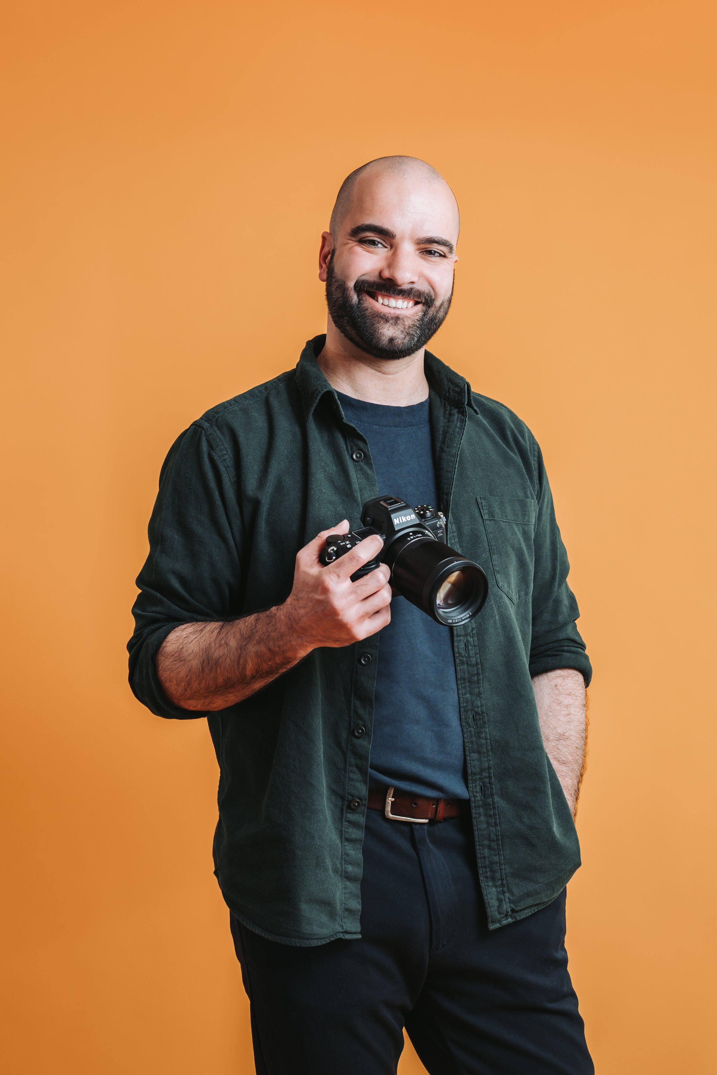Ein Porträt von Hochzeitsfotograf Dominic Schleicher-Glock, dem Mann hinter der Kamera von Dominic Glock Fotografie. Er trägt einen kurzen Bart und Glatze. Er hält eine Nikon-Kamera und lächelt vor einem orangefarbenen Hintergrund.