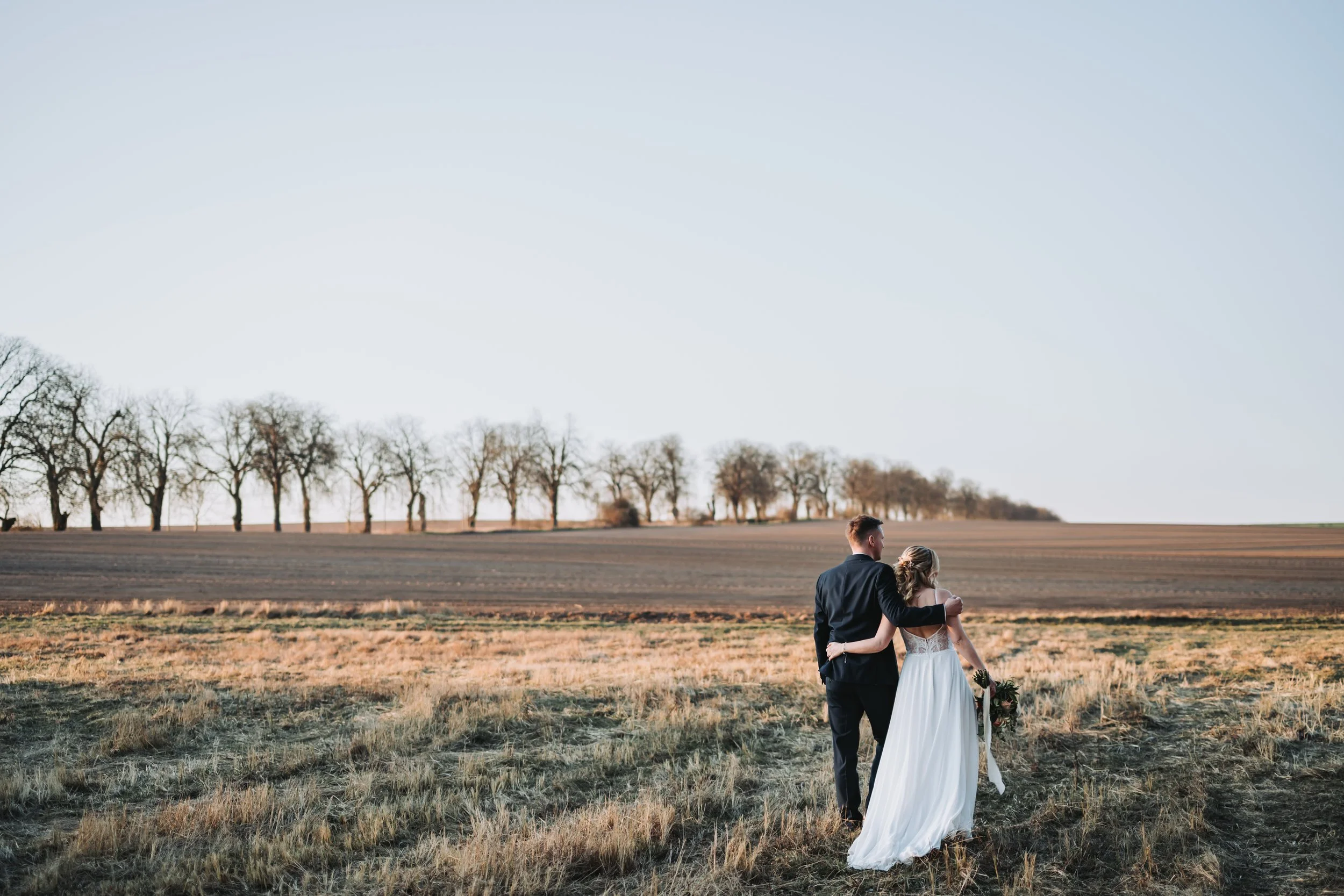 Ein Brautpaar spaziert gemeinsam über eine offene Wiese mit einem Baumreihe im Hintergrund bei Sonnenuntergang in der Natur. Aufgenommen von Hochzeitsfotograf Dominic Glock.