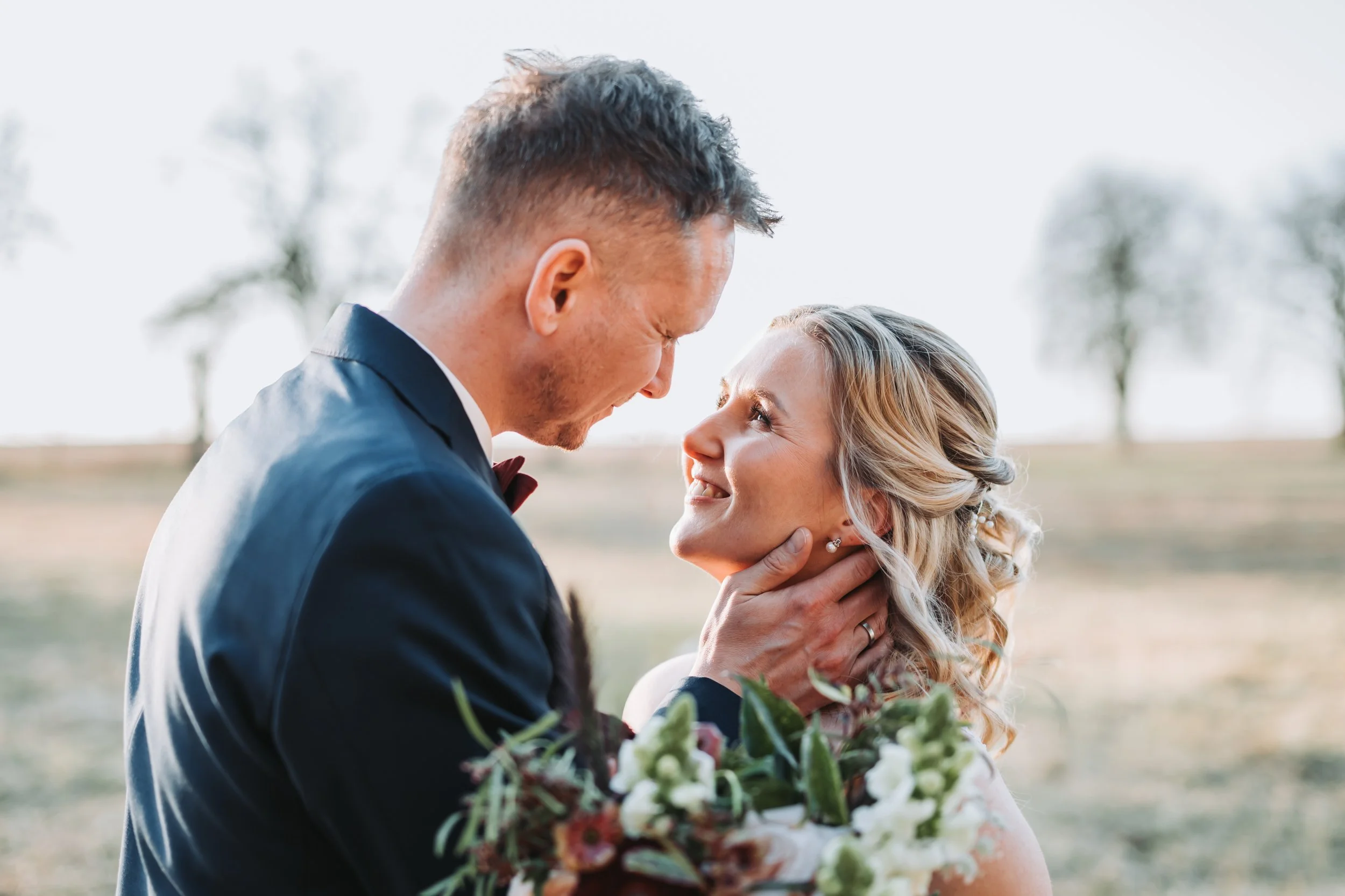 Ein Brautpaar bei einem Hochzeitsshooting im Freien, der Mann hält die Frau liebevoll im Arm, beide lächeln sich verbunden an, im Hintergrund sind Bäume und ein klarer Himmel.