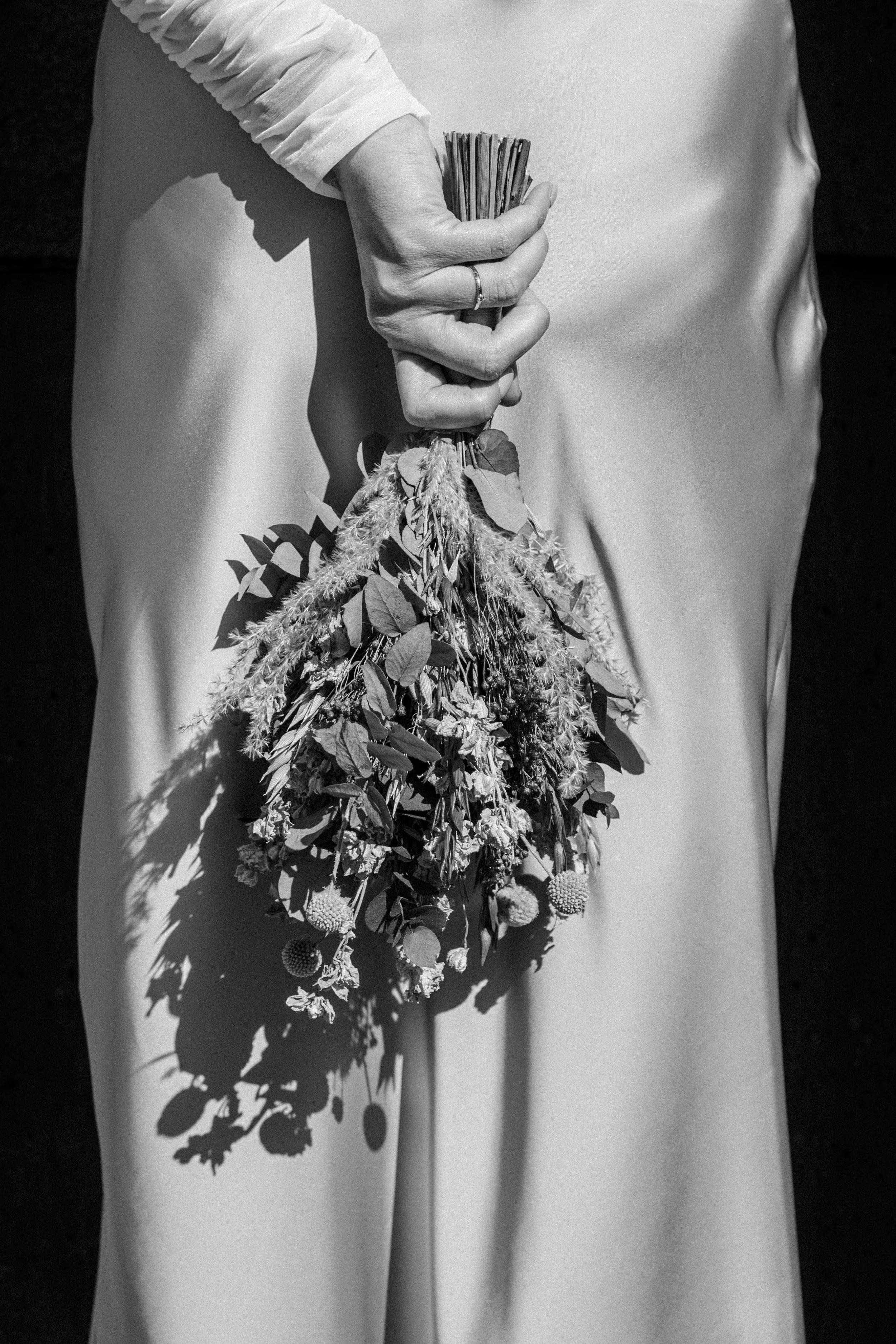 Schwarze und weiße Fotografie Braut mit einer Hand, die einen Brautstrauß aus getrockneten Pflanzen und Blumen hält, vor einem satinierten Kleid oder Stoff, Schatten wirft.