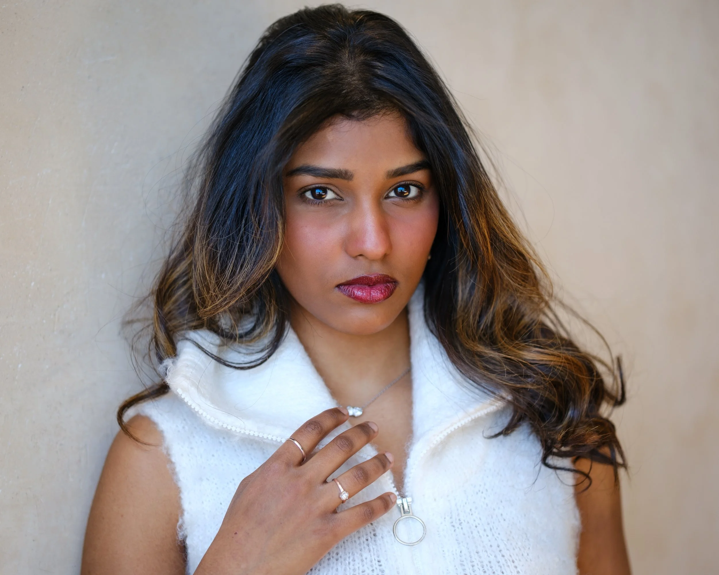 Porträt einer jungen Frau mit dunklen, gewellten Haaren, die einen weißen, ärmellosen Pullover trägt, vor einer hellen Wand, Blick direkt in die Kamera, mit Schmuck an Hals, Finger und Ohren.