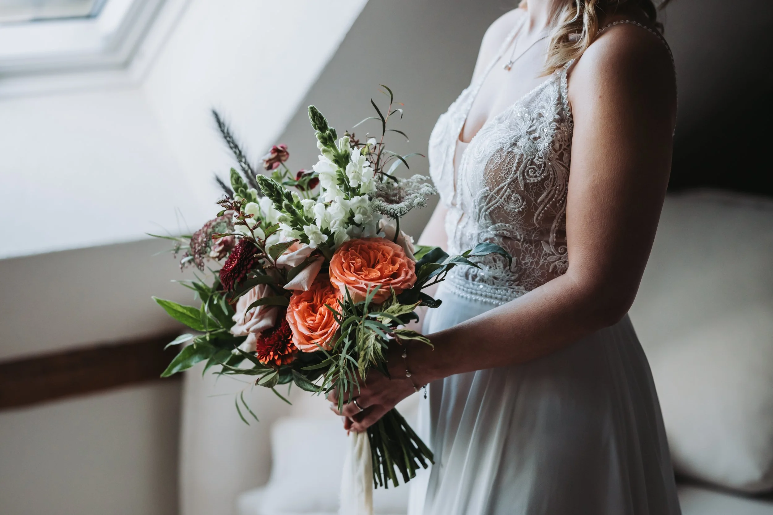 Eine Frau in einem weißen Hochzeitskleid hält einen Blumenstrauß mit weißen, orangefarbenen und roten Blumen vor einer Fensterseite.