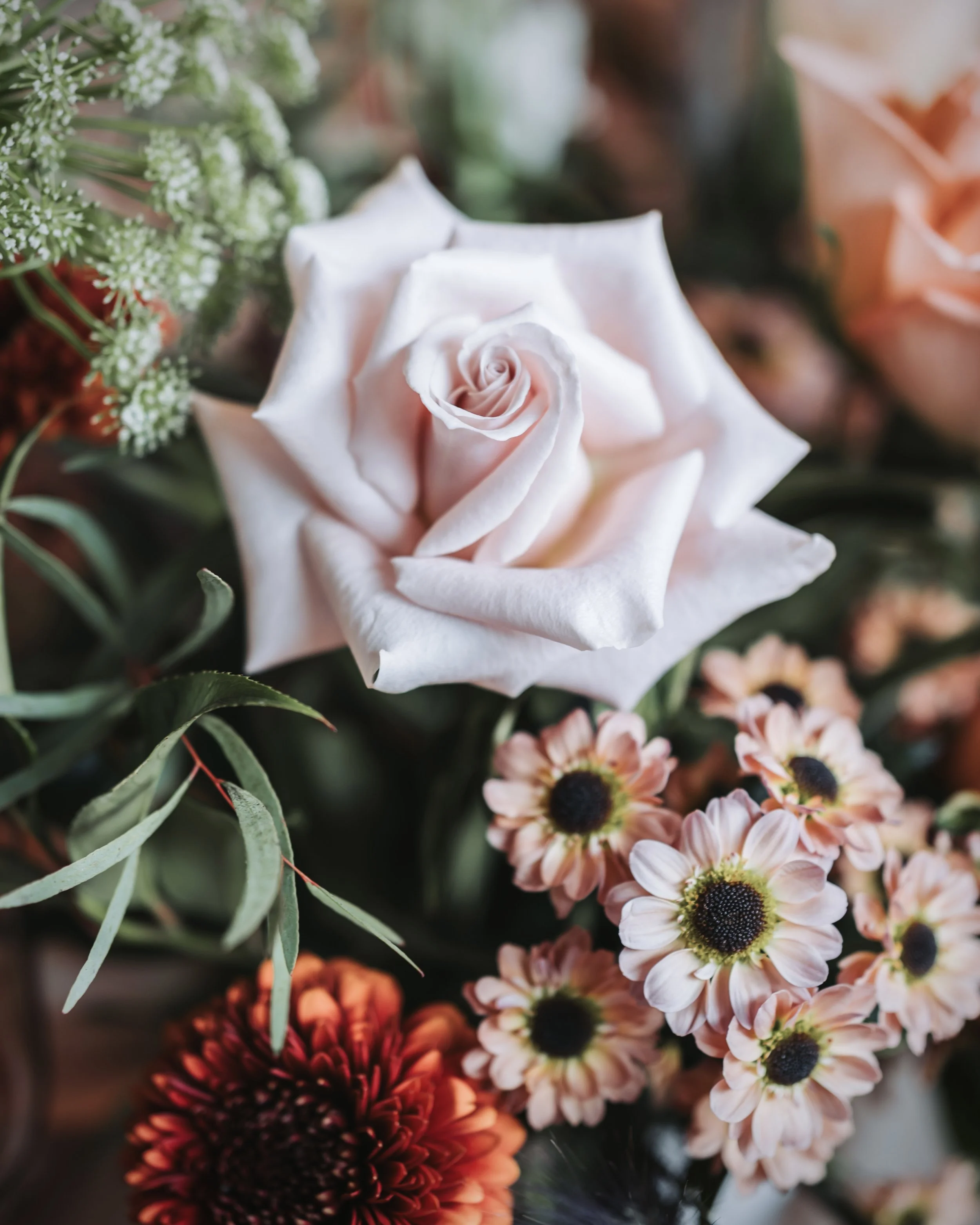 Eine weiße Rose inmitten verschiedener anderer Blumen, darunter kleine rosa Chrysanthen und grüne Blätter. Aufgenommen von Hochzeitsfotograf Dominic Glock.