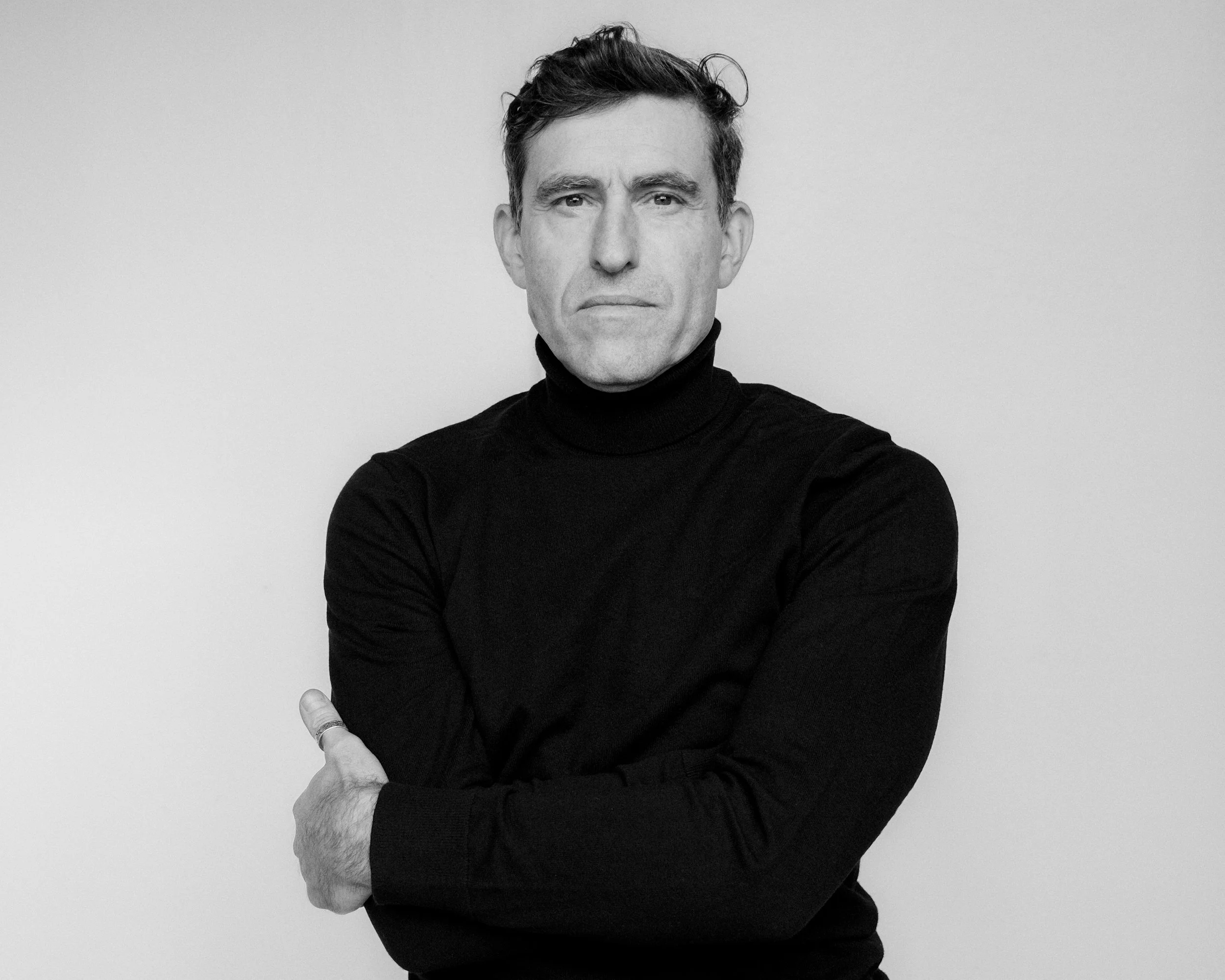 Schwarz-weiß-Porträt eines Mannes in einem schwarzen Rollkragenpullover, mit verschränkten Armen, frontale Perspektive, neutraler Gesichtsausdruck, einfacher heller Hintergrund.