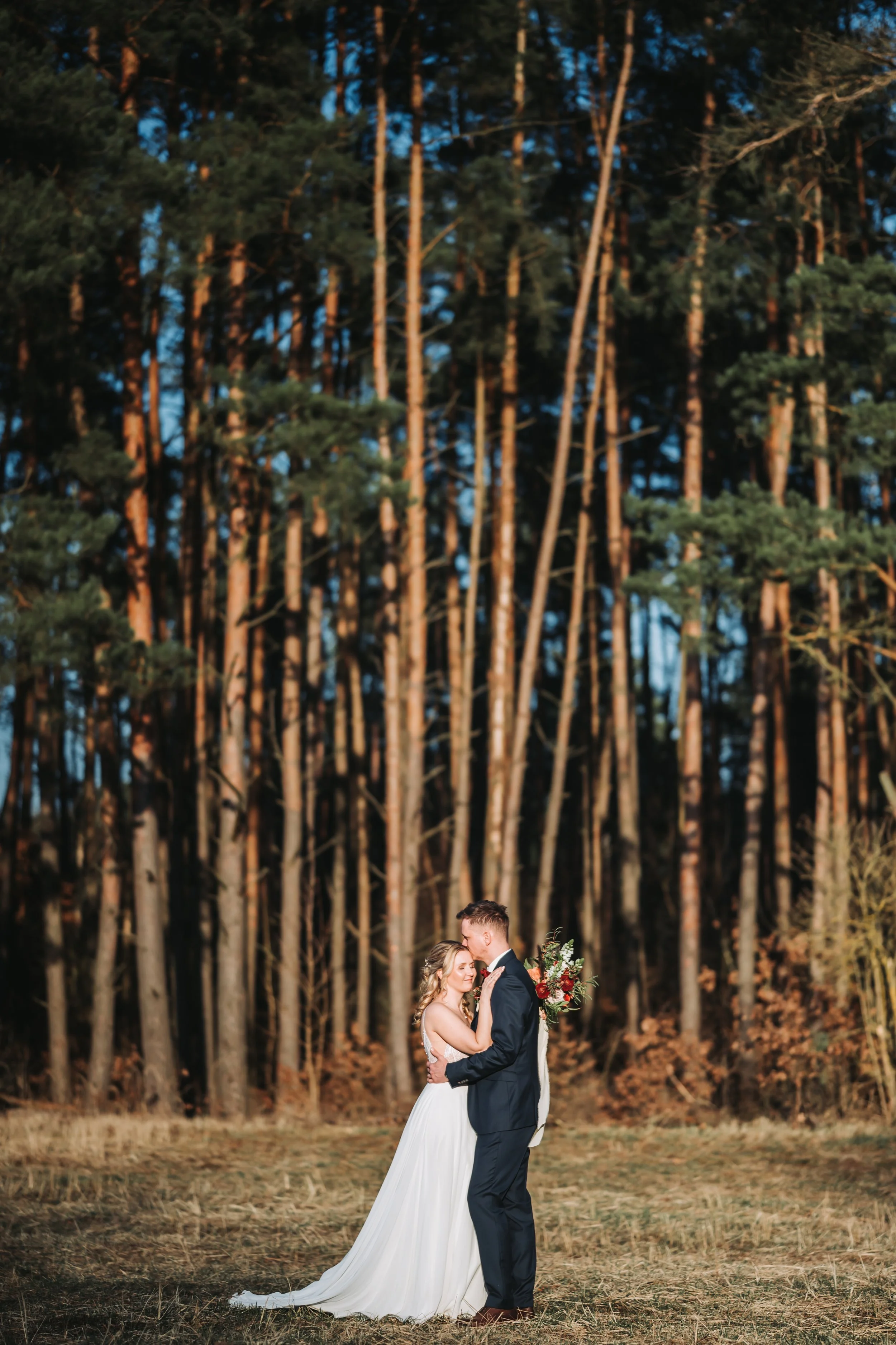 Ein Brautpaar umarmt sich in einem Wald bei Sonnenuntergang, die Braut trägt ein weißes Hochzeitskleid und der Bräutigam einen dunklen Anzug, hält einen Blumenstrauß. Aufgenommen von Hochzeitsfotograf Dominic Glock.