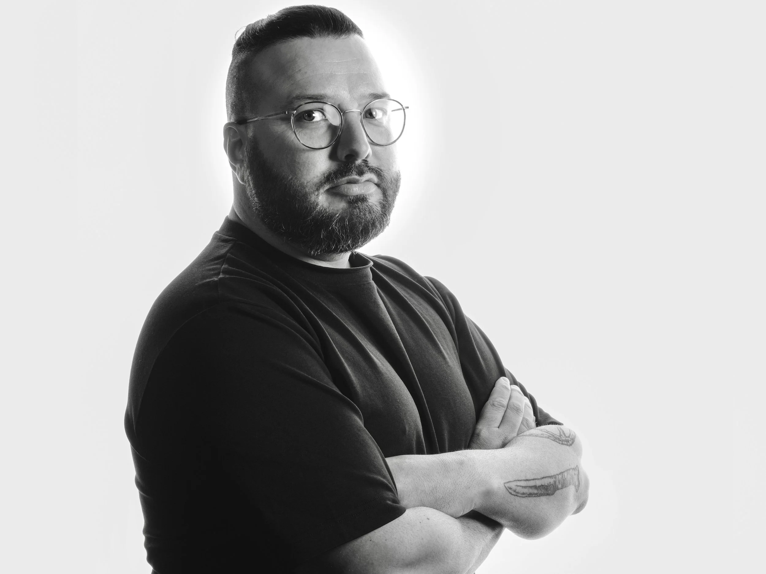 Schwarz-weiß Porträt eines Mannes mit Brille, kurzem Haar und Bart, der die Arme verschränkt trägt, auf hellem Hintergrund.