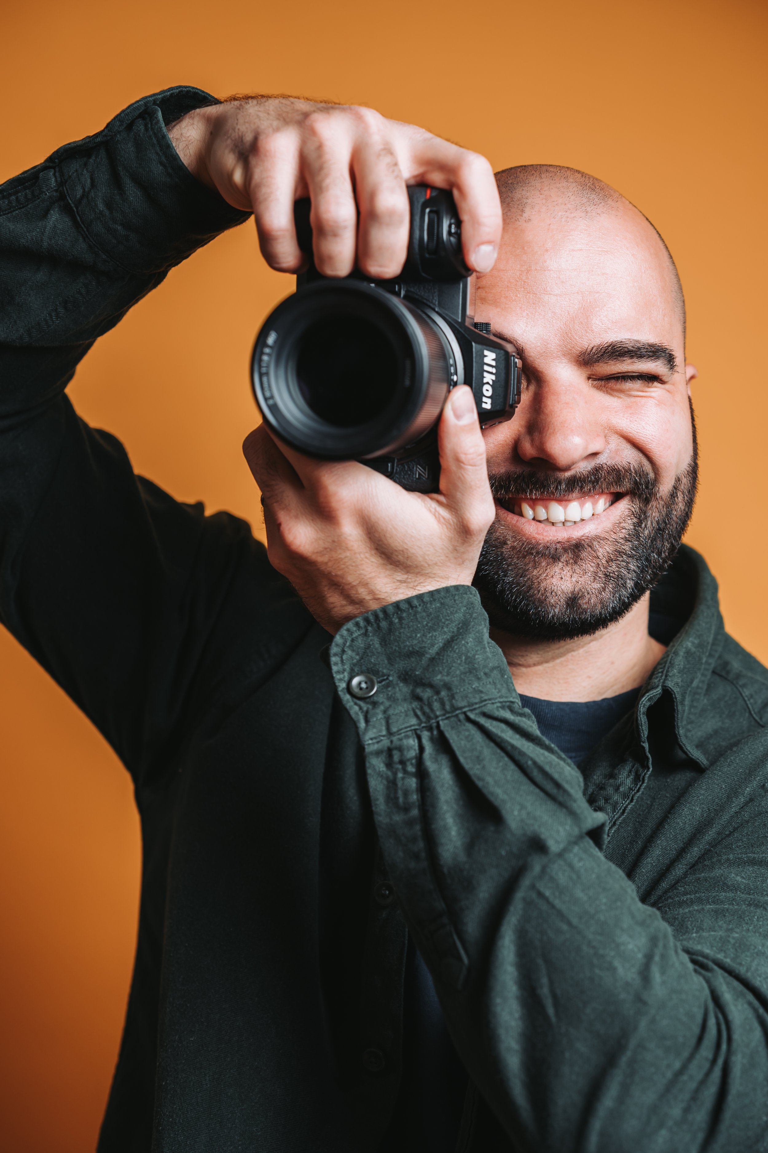 Ein Porträt von Hochzeitsfotograf Dominic Schleicher-Glock, dem Mann hinter der Kamera von Dominic Glock Fotografie. Er hält eine Nikon-Kamera vor sein Auge und lacht dahinter, vor einem orangefarbenen Hintergrund.