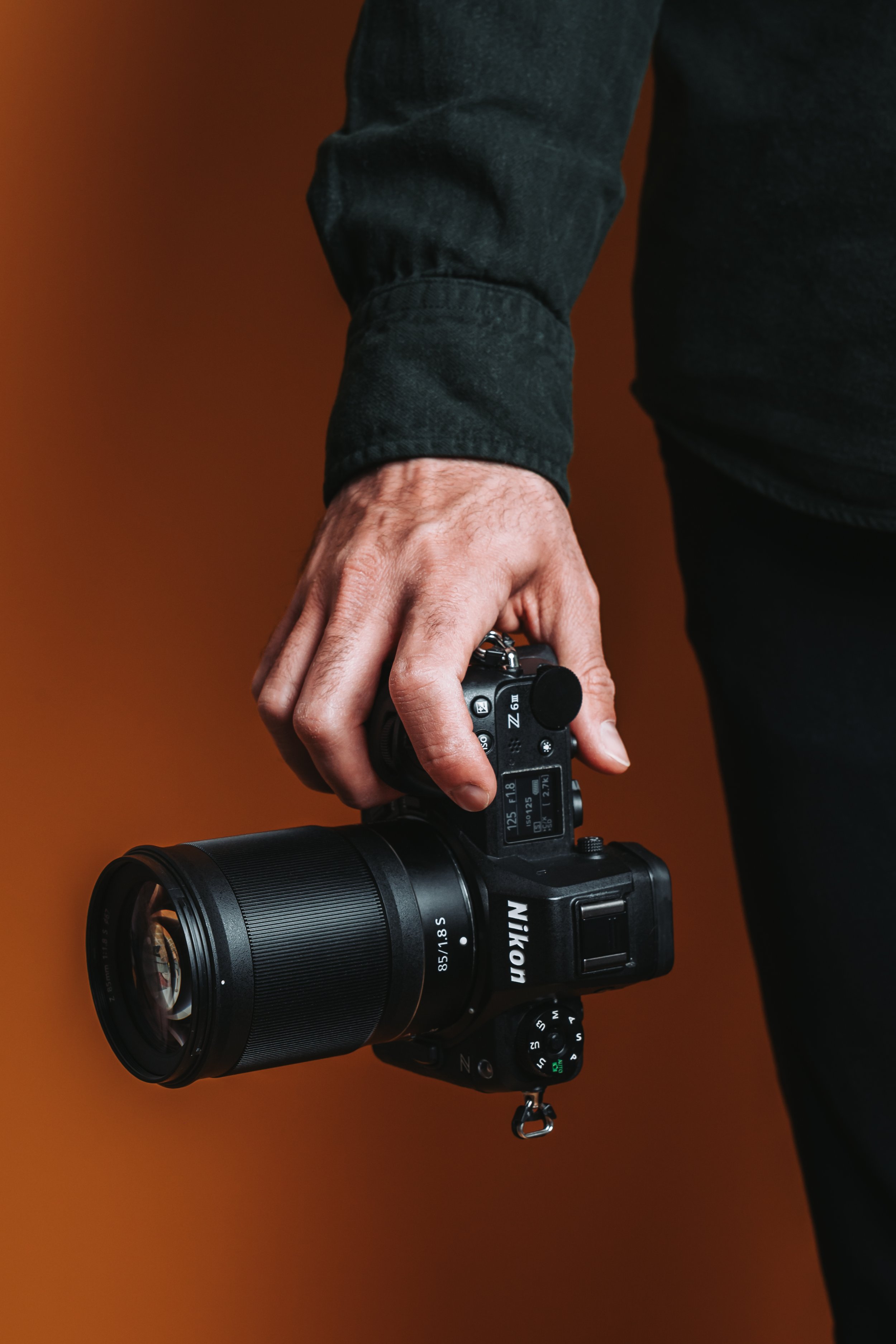 Die Hand von Hochzeitsfotograf Dominic Schleicher-Glock hält eine Nikon-Kamera mit großem Objektiv vor einem dunkel orangenen Hintergrund.