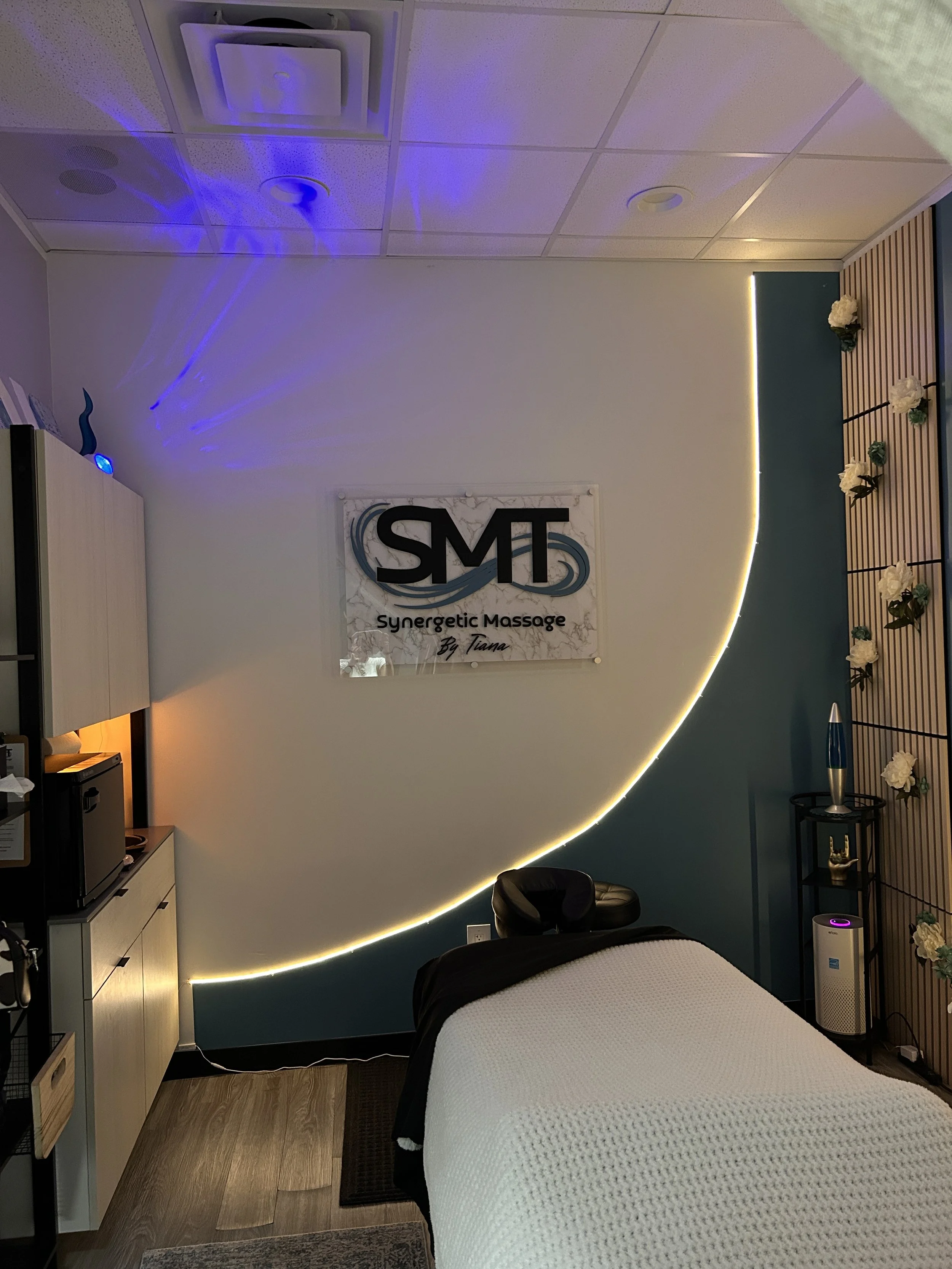 A massage room with a bed covered in a plush beige blanket, a small shelf with a lava lamp, a diffuser, and decorative flowers, illuminated by warm lighting and wall art featuring text.