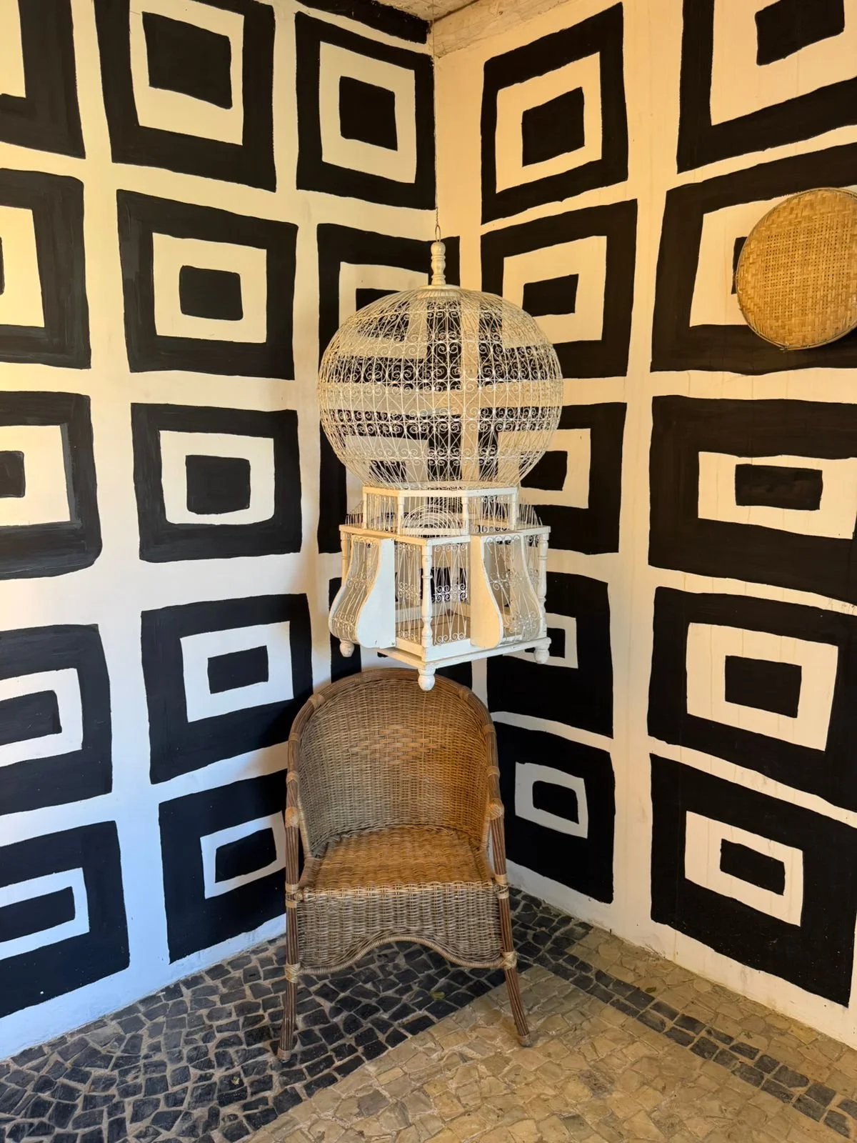 A corner of a room with black and white geometric patterned walls, a wicker chair, a white birdcage-style light fixture hanging from the ceiling, and round wicker wall decor.