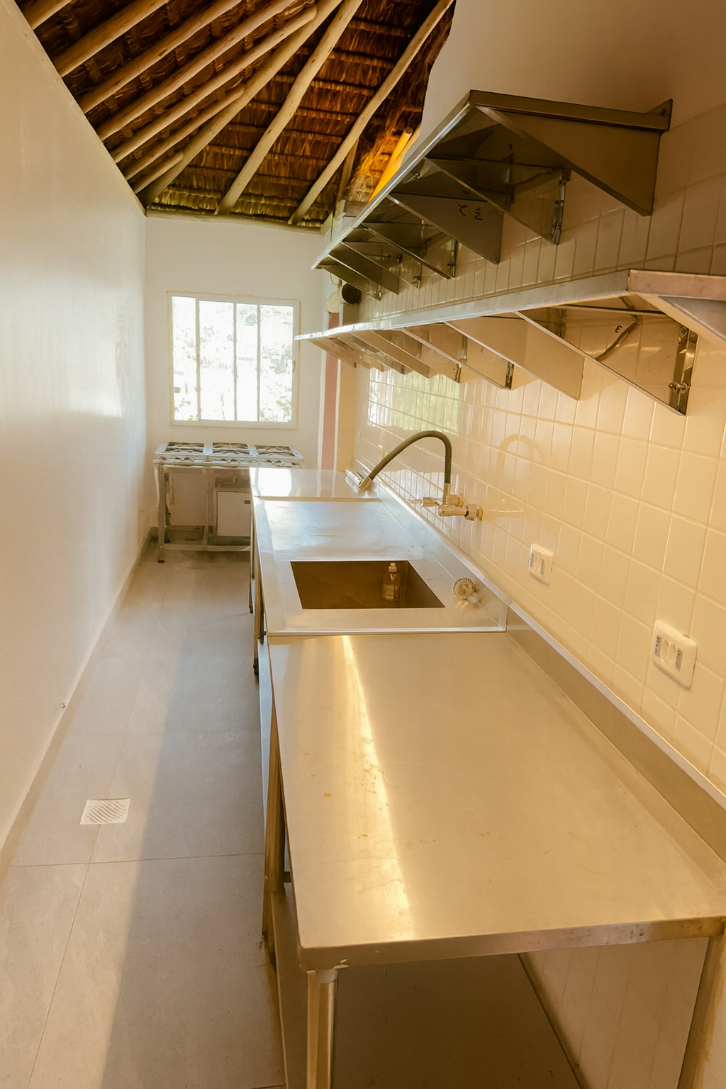 Empty commercial kitchen with stainless steel counters, a sink, and wall-mounted exhaust hoods, natural light coming through a window, and a tiled wall.