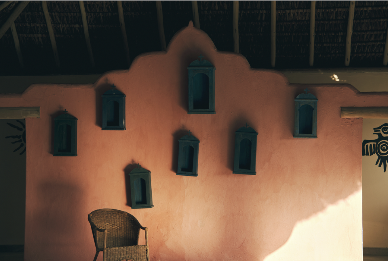 A pink stucco wall with seven blue shadow boxes and a wicker chair in the foreground, under a thatched roof ceiling.