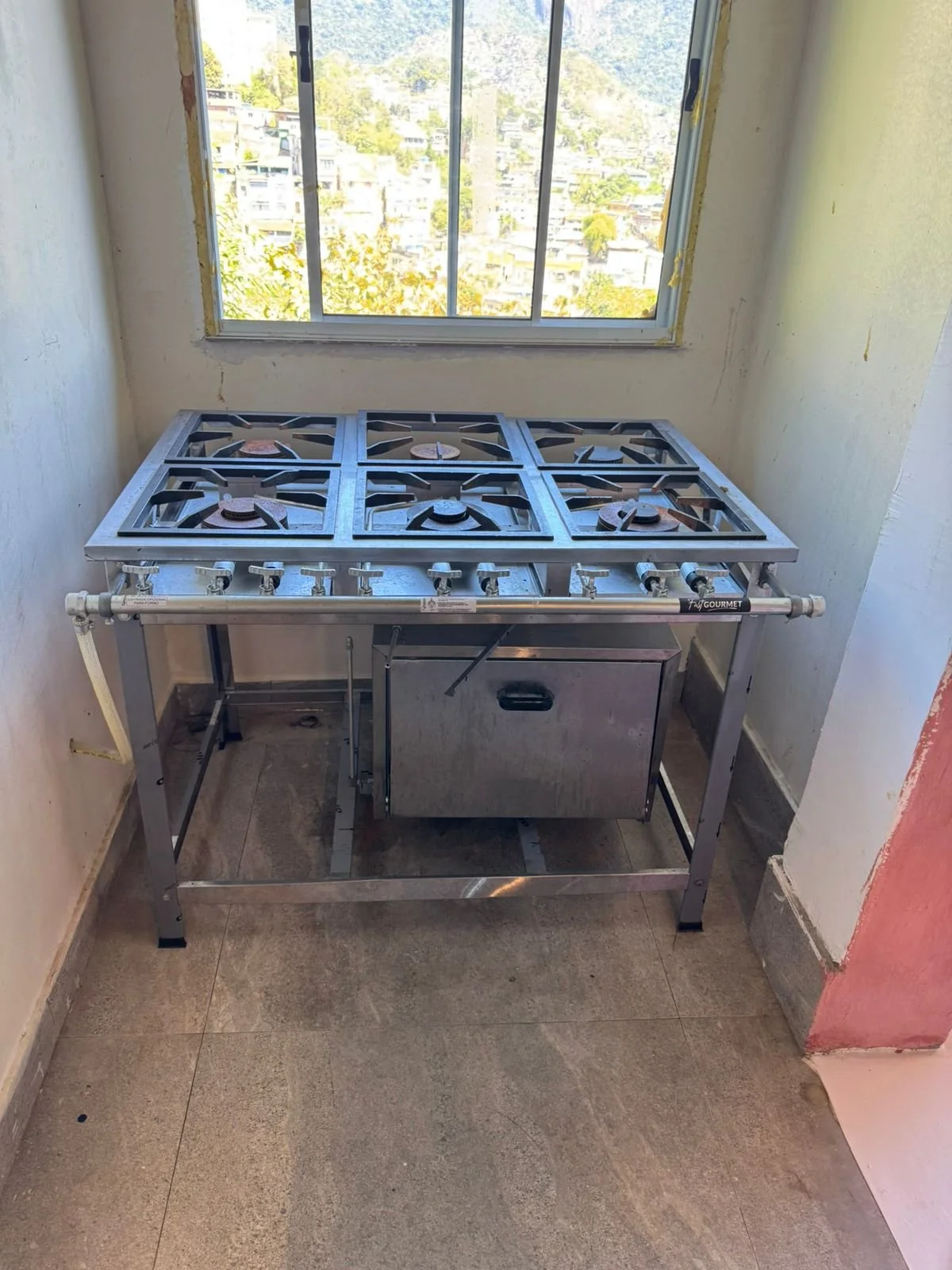 A stainless steel six-burner gas stove with a storage drawer underneath, situated in a small, unfinished room with a window showing a view of a hillside neighborhood.