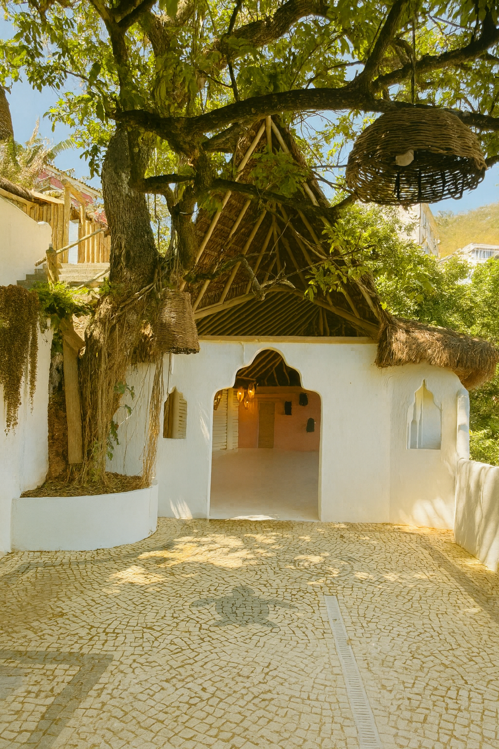 A small white building with an arched entrance, surrounded by a courtyard with cobblestone paving. There are trees with green leaves growing over the roof, and woven baskets hanging from the branches. The building's roof is made of thatched straw, and the sky is blue with sunlight filtering through the leaves.