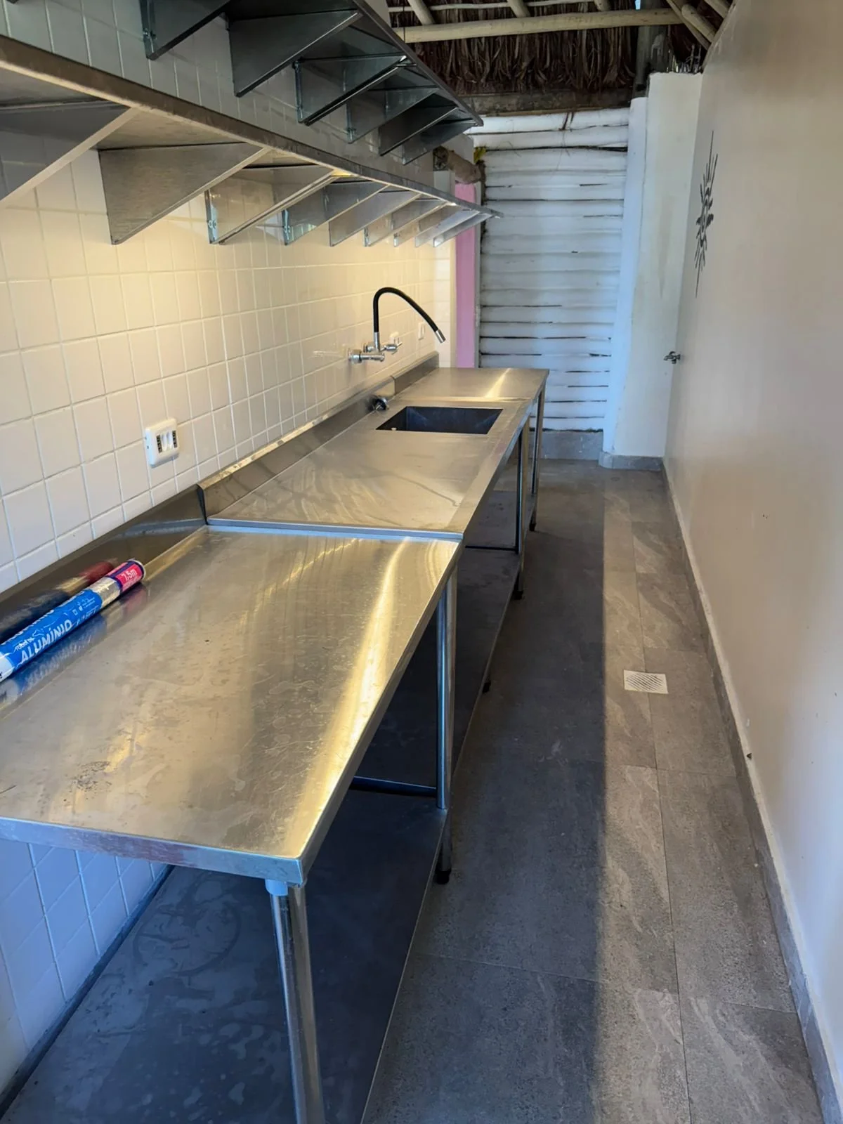 Stainless steel countertop with a single basin sink, a black faucet, and overhead shelves, in a narrow kitchen or utility space with tiled wall and a white wooden fence at the back.