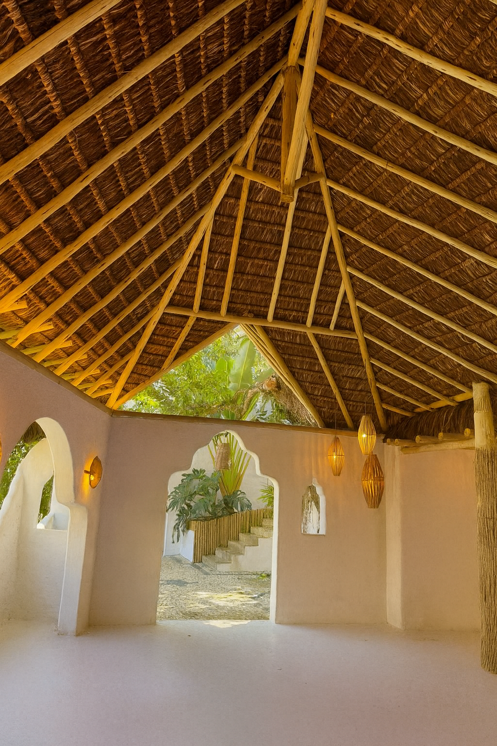 Interior view of a thatched roof ceiling with wooden beams, decorative wall niches, and hanging lanterns, with an open archway leading to an outdoor area with stairs and lush greenery.