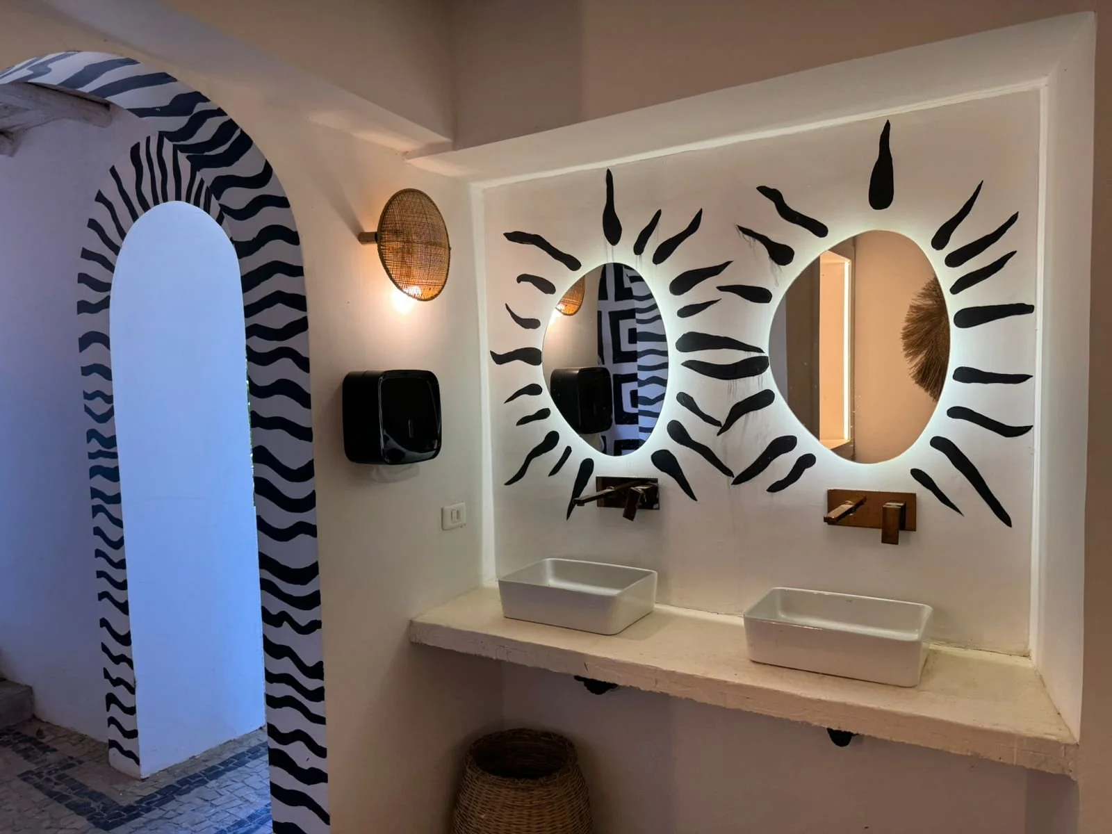 A bathroom with two white rectangular sinks on a counter, two round mirrors with decorative designs radiating from their edges, and black wall-mounted soap dispensers. The wall has black and white zebra stripe accents around the arched doorway and mirrors, with wall-mounted light fixtures.