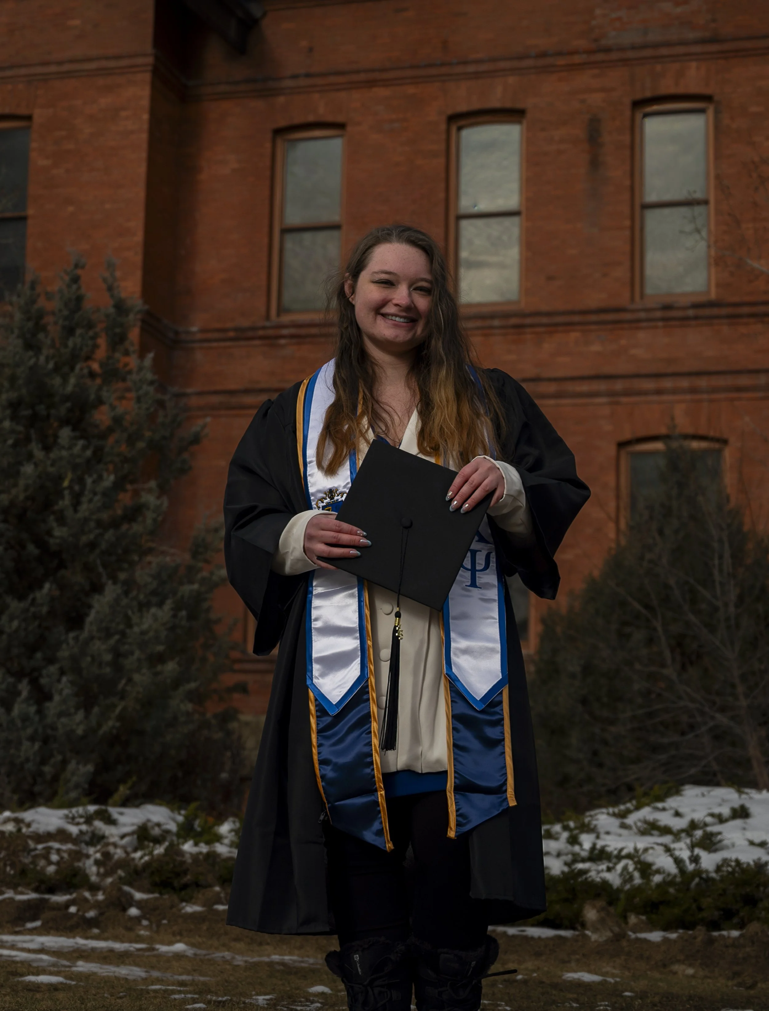 A young woman in graduation gown and cap holding diploma, standing outside in front of a brick building with snow on the ground.