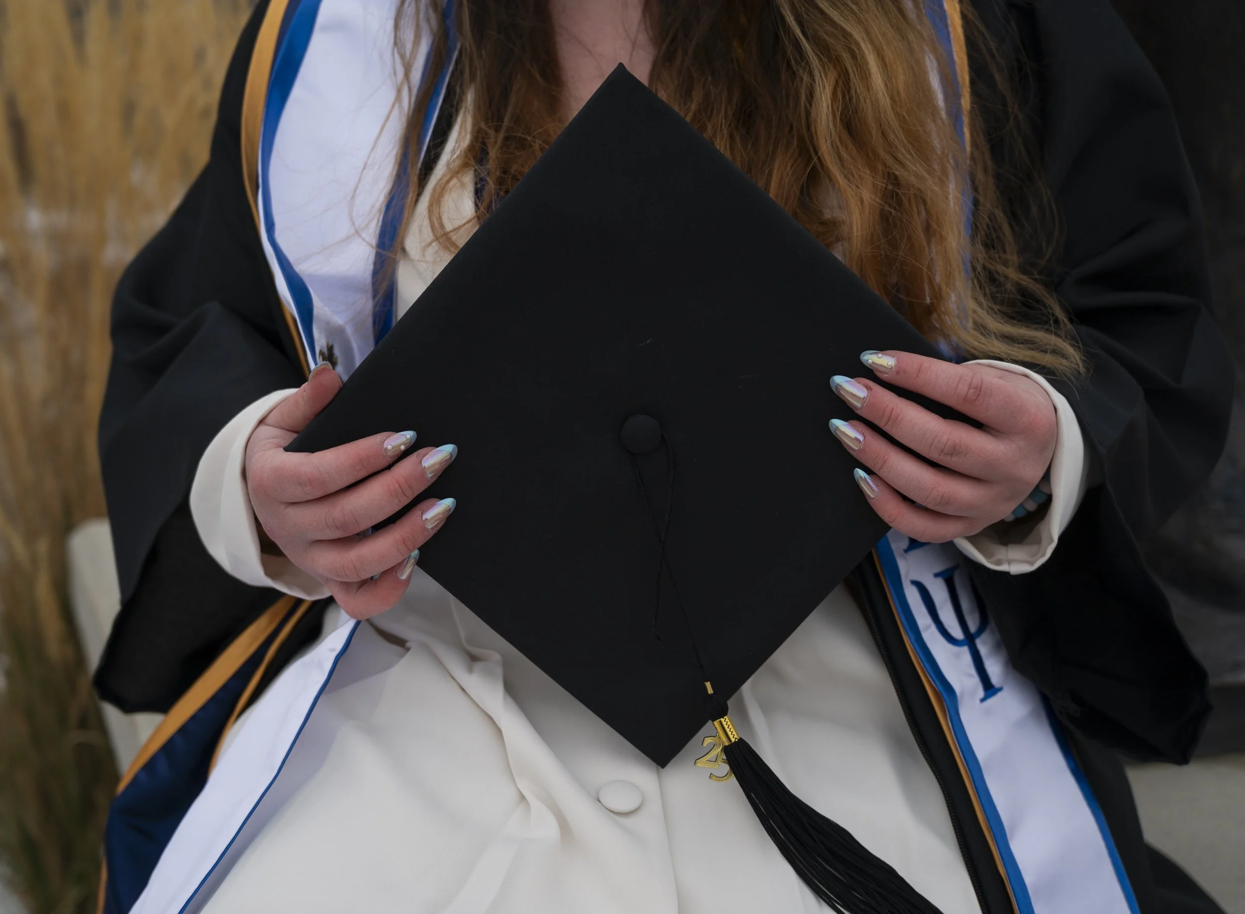 Close-up of a person holding a graduation cap with a tassel, wearing a graduation gown and a sash.