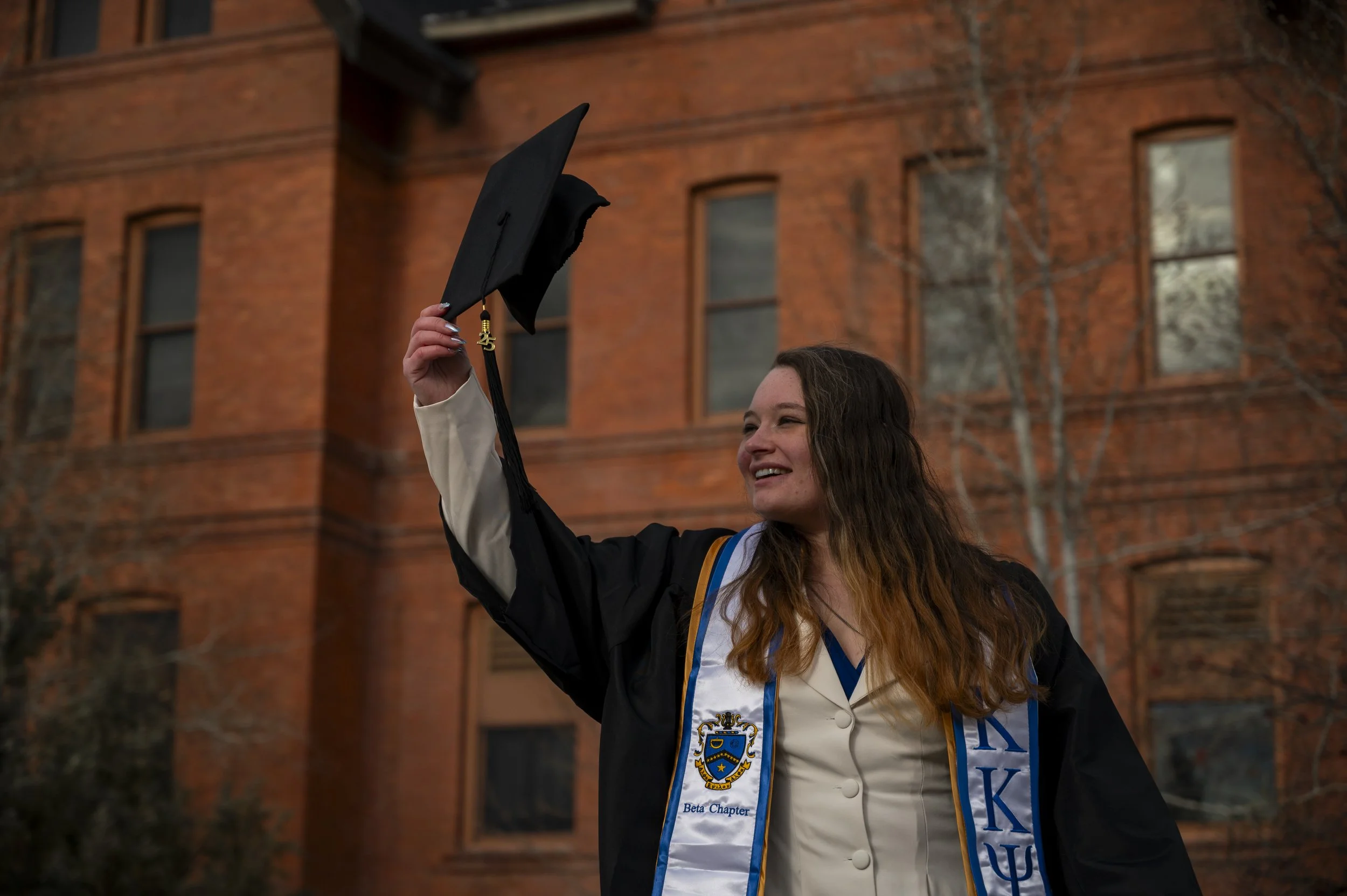 Young woman in graduation cap and gown holding her cap in the air, smiling, standing outside in front of a brick building.