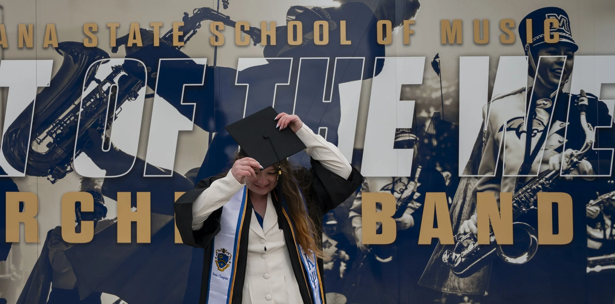 A young woman in graduation cap and gown smiling as she adjusts her cap, standing in front of a mural with a saxophone-playing band at a music school.