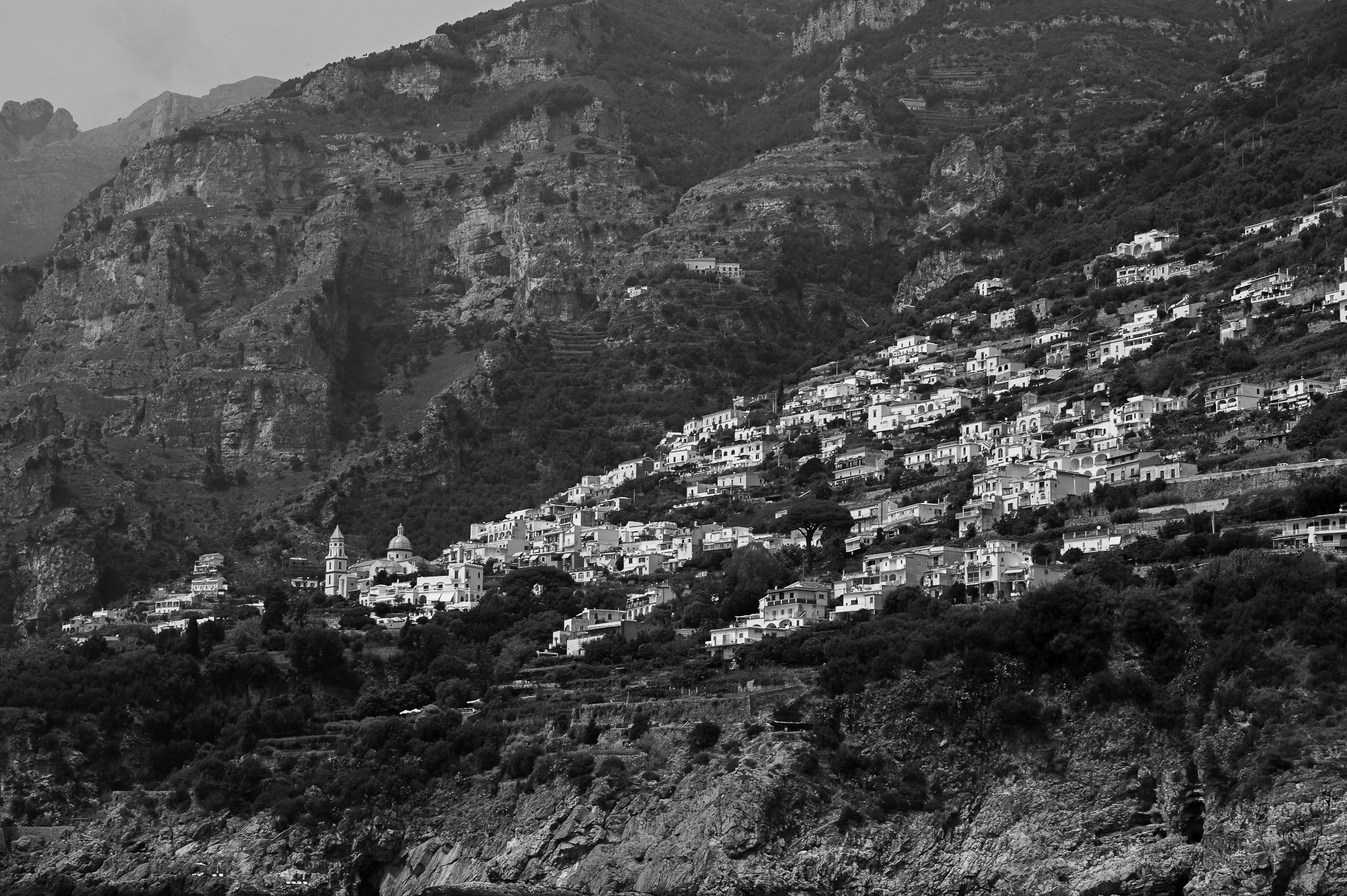 A black and white photo of a steep hillside with densely packed white houses and a church with a tall bell tower, set against rocky mountains.