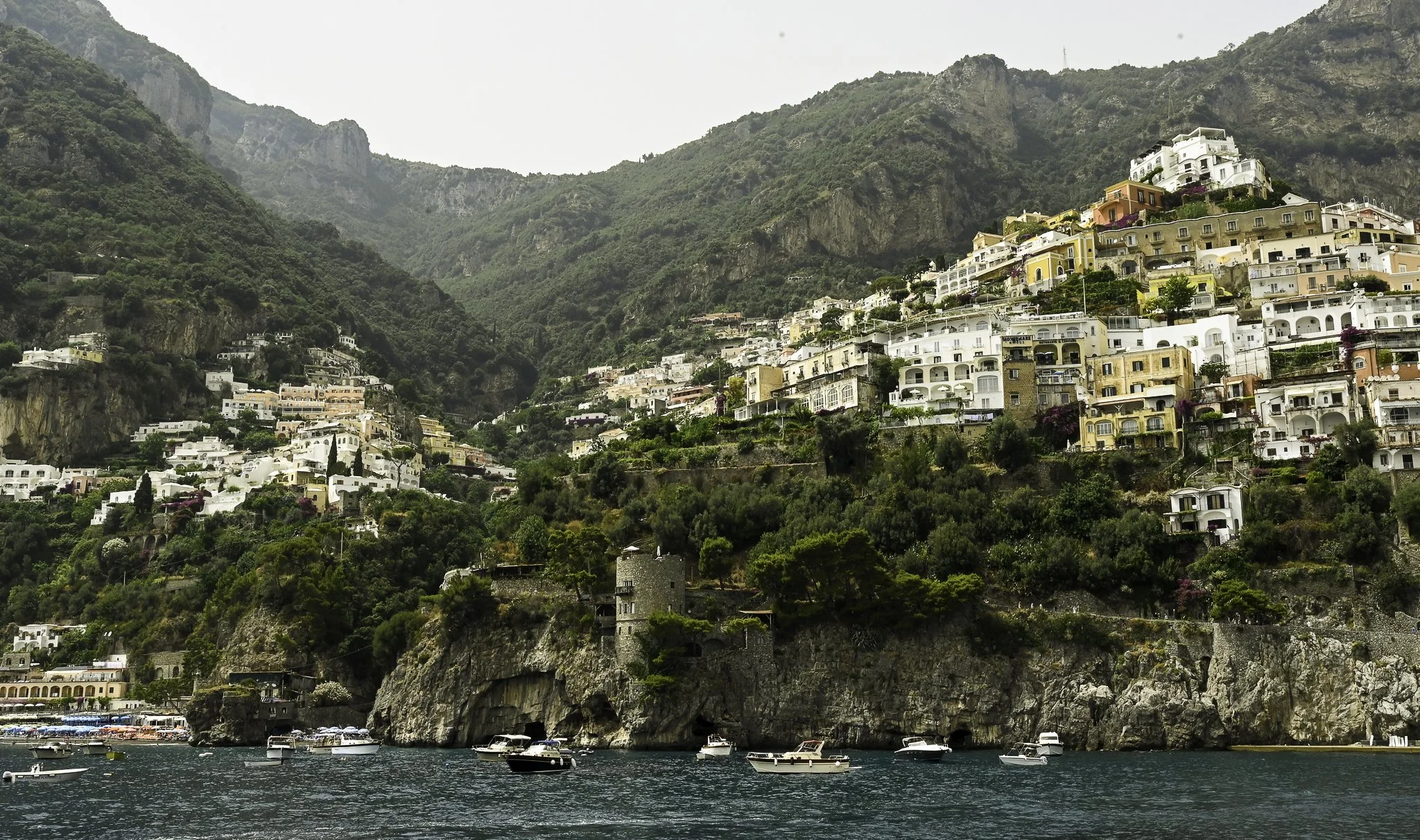 Hillside town with colorful houses above water, boats docked at shoreline, and rugged mountainous background.