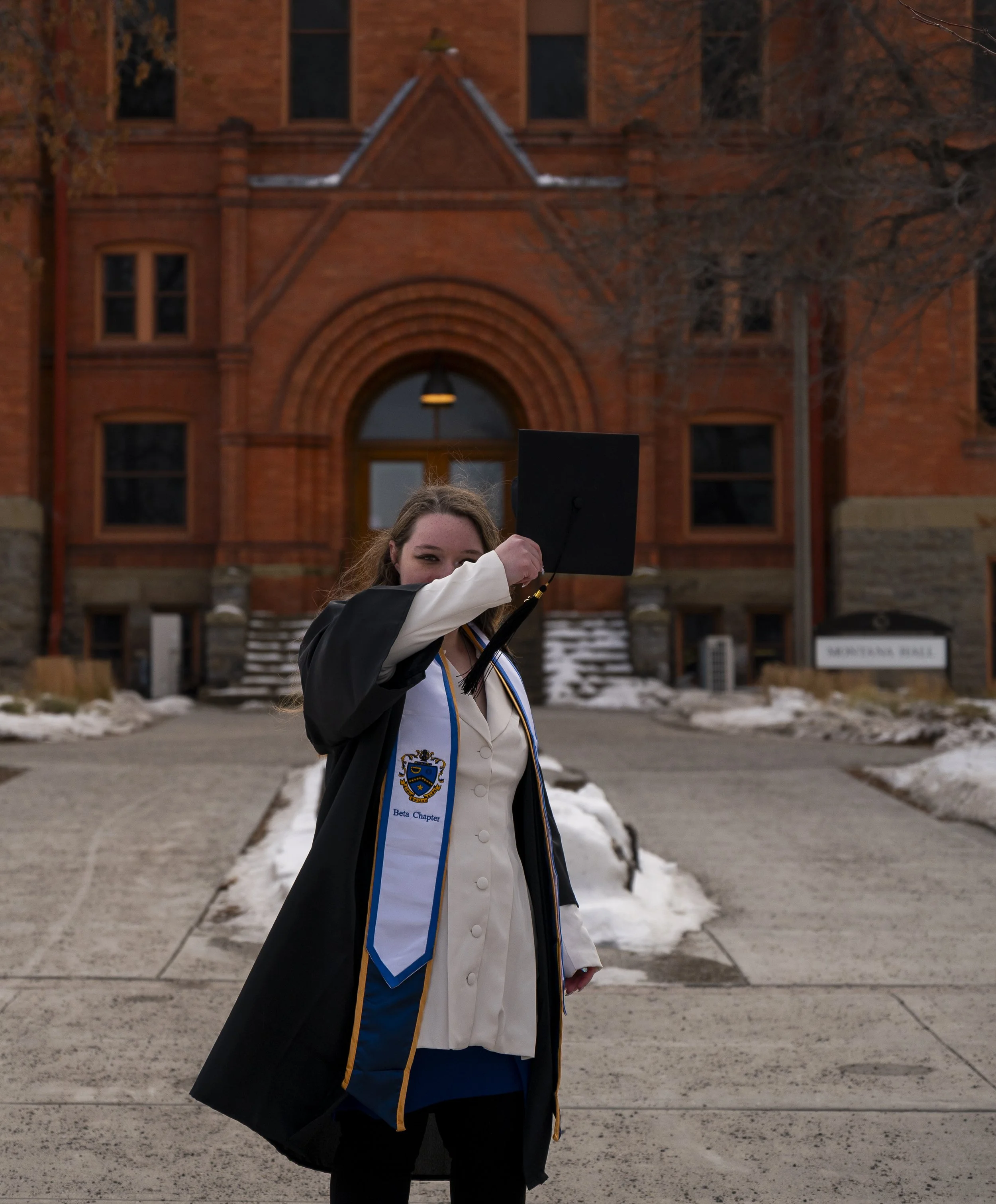 A young woman in graduation gown holding her cap up on a college campus with a brick building and snow on the ground.