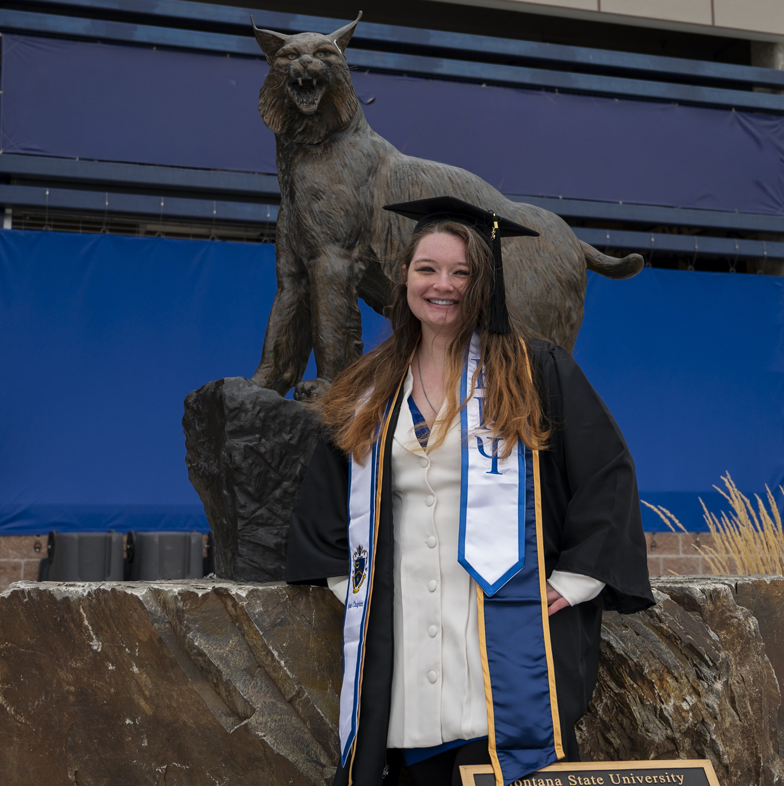 A smiling woman in graduation attire, including a cap and gown, standing in front of a bronze sculpture of a bobcat and a cougar at Montana State University.