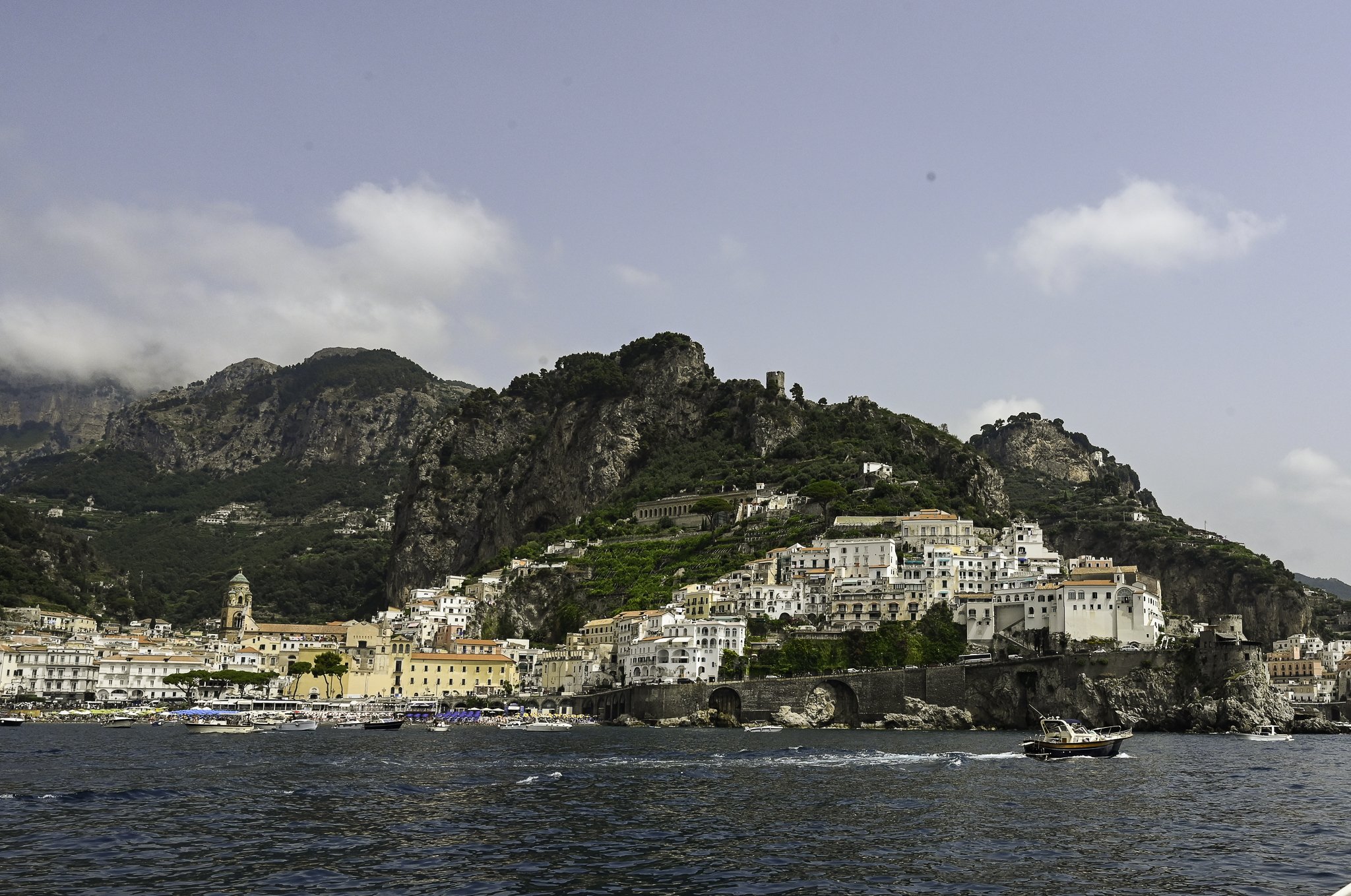 View of a coastal town with white buildings on a hillside, boats in the water, and mountains in the background.