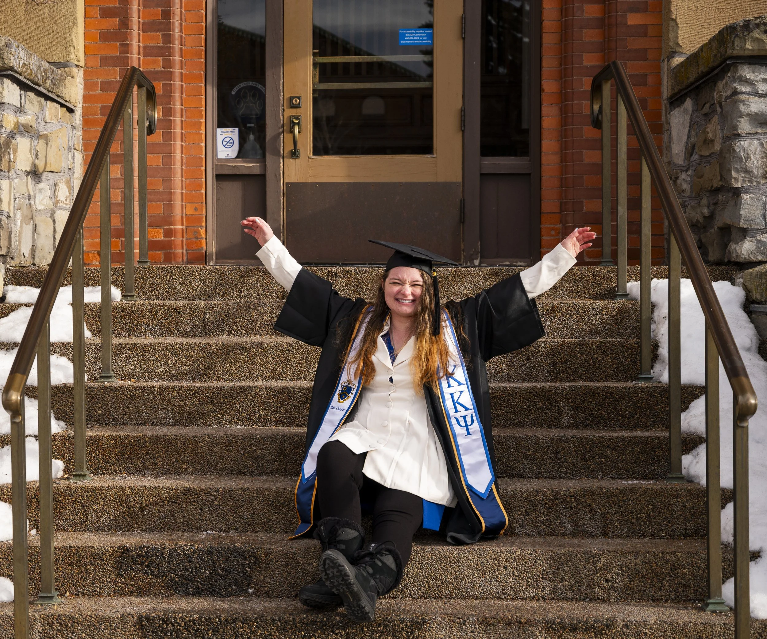 A young woman in graduation cap and gown celebrating on the steps outside a building, smiling with her arms raised.
