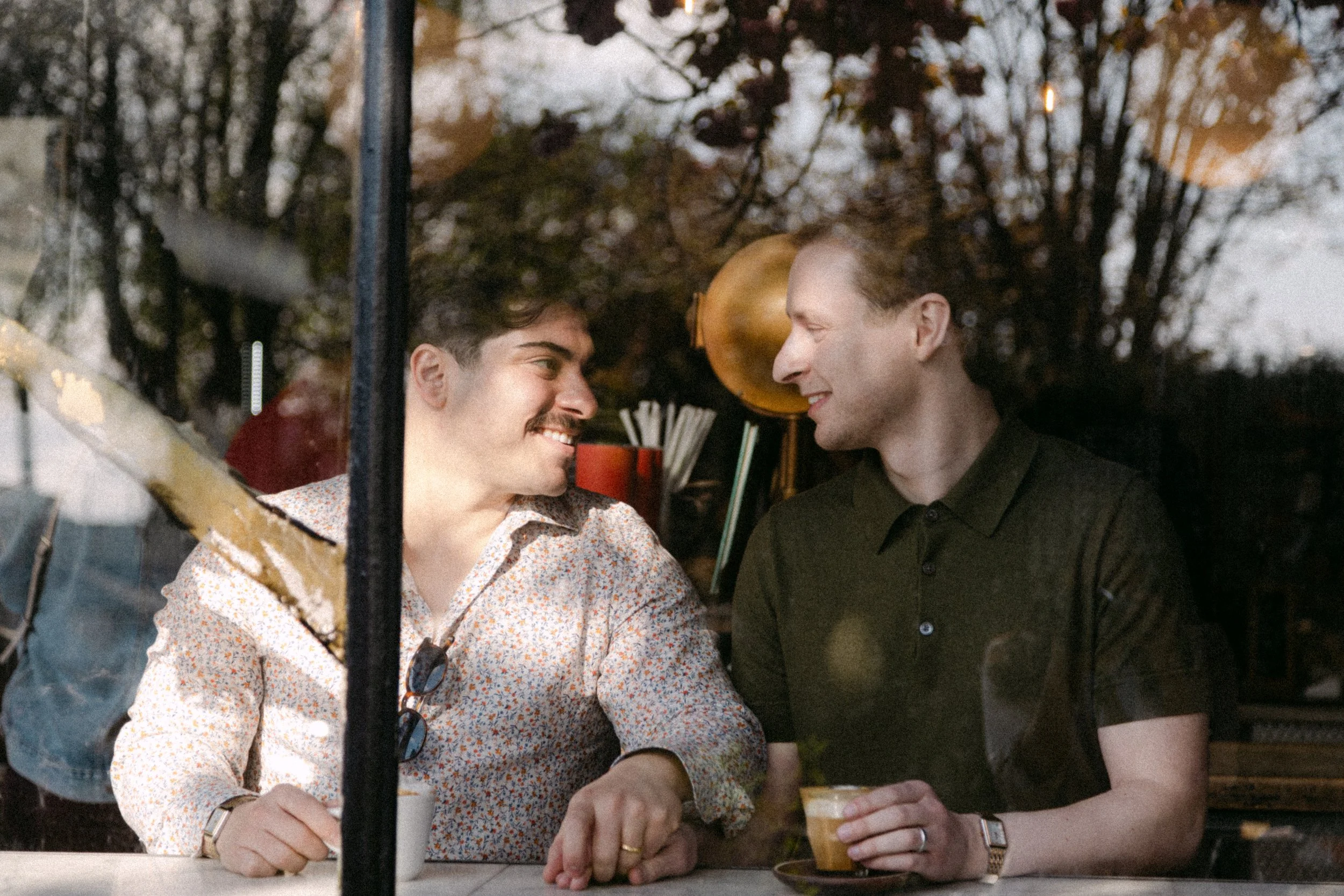 Two men sit at a table outside, smiling and looking into each other's eyes, in a cozy, outdoor setting during the evening.