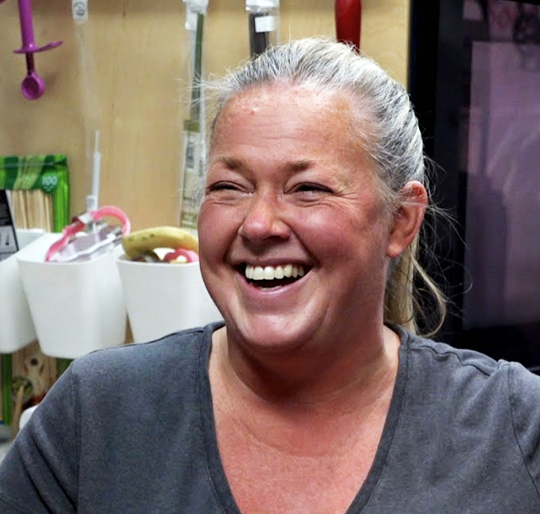 A woman with blonde hair tied back, smiling and laughing in what appears to be a kitchen or workspace.