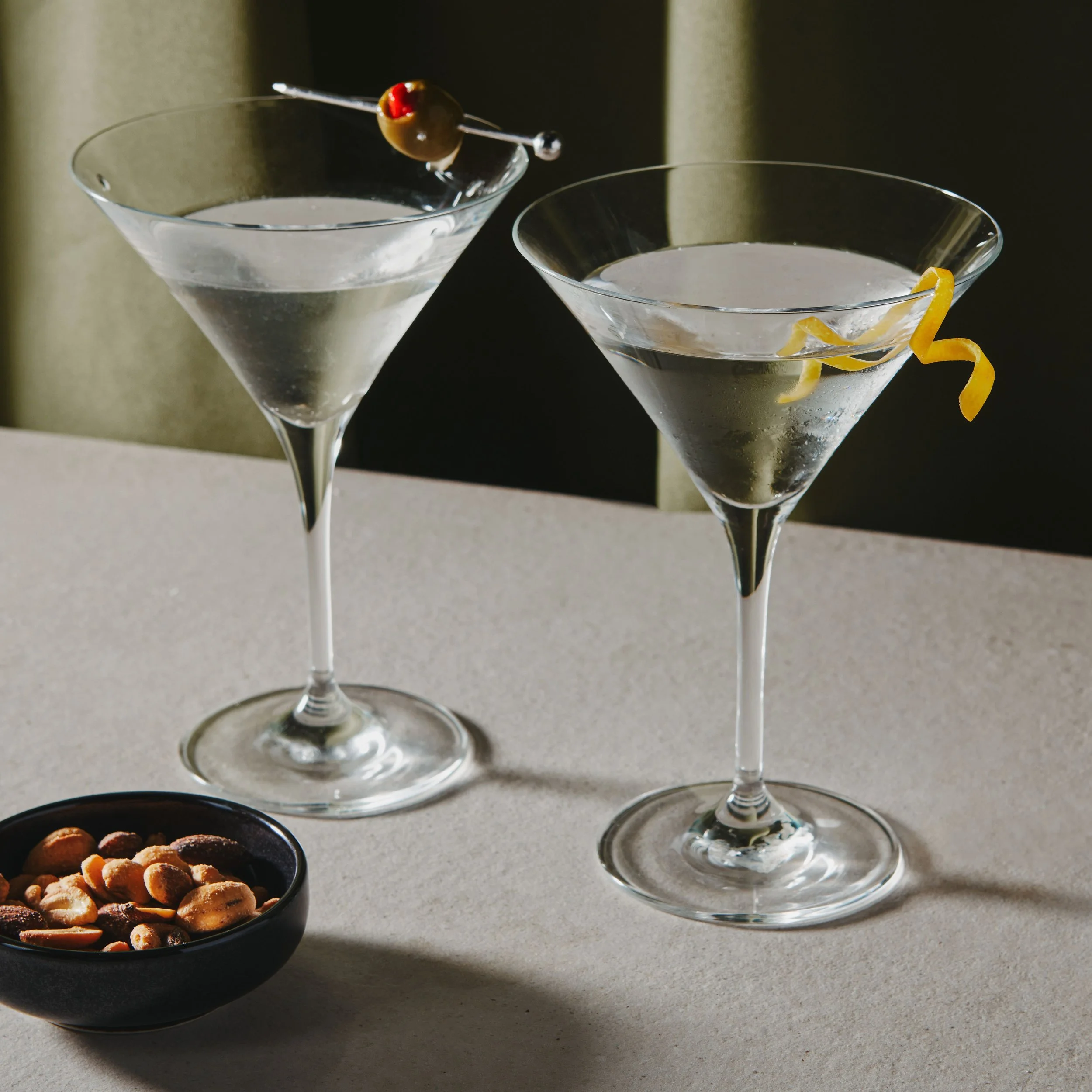 Two martini glasses with clear liquid, garnished with a lemon twist and an olive skewer, placed on a light-colored surface with a bowl of mixed nuts nearby.