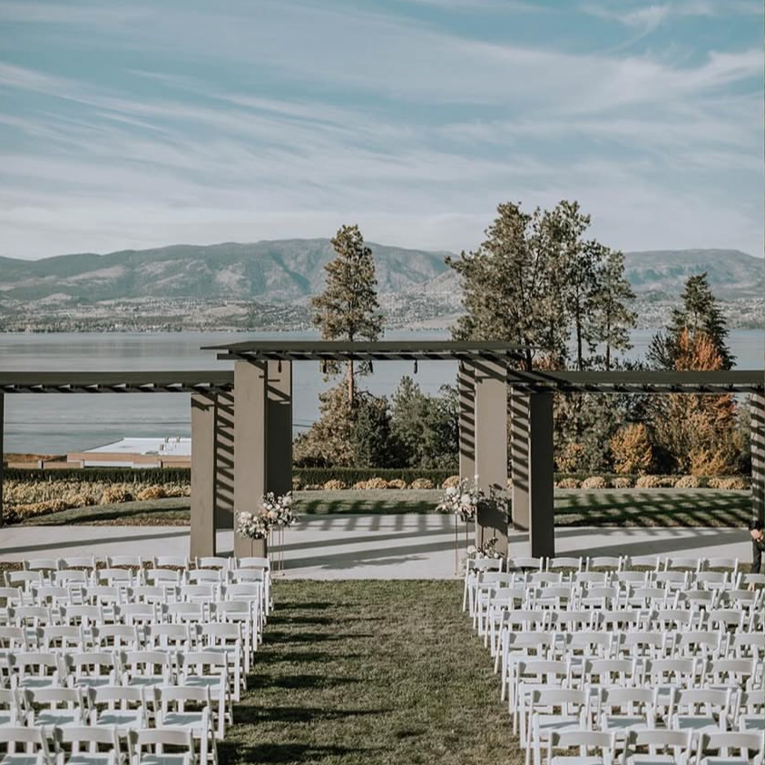 Outdoor wedding ceremony setup with white chairs on either side of a grassy aisle, floral arrangements, and a scenic backdrop of a lake, trees, and mountains under a partly cloudy sky.