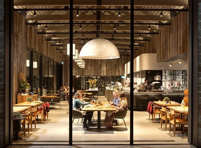 Inside a modern restaurant with several patrons dining, wooden walls, modern lighting, and an open kitchen area in the background.