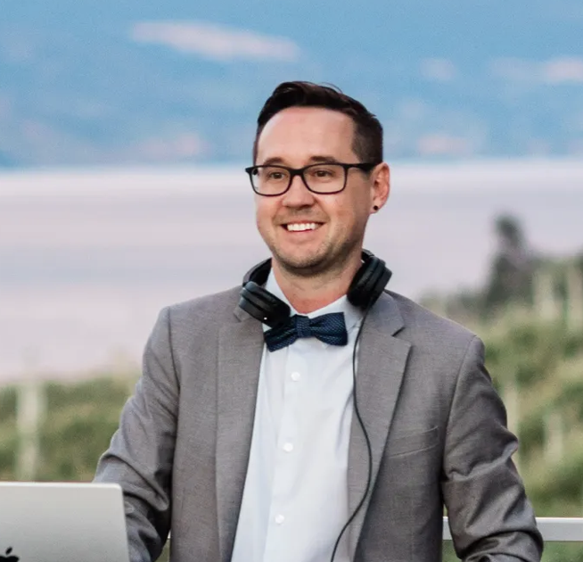 A man with glasses, wearing a gray suit and bow tie, smiling outdoors with a blurred landscape and blue sky in the background.