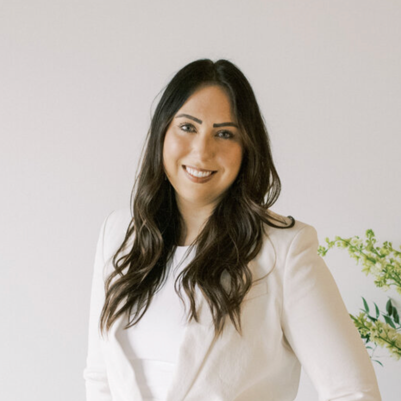 A woman with long dark hair, wearing a white blazer, smiling at the camera against a plain background with some green plants.