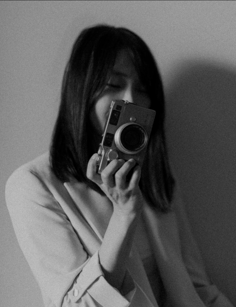 Black and white photo of a woman taking a selfie with a camera, with her eyes closed and a relaxed expression, against a plain background.