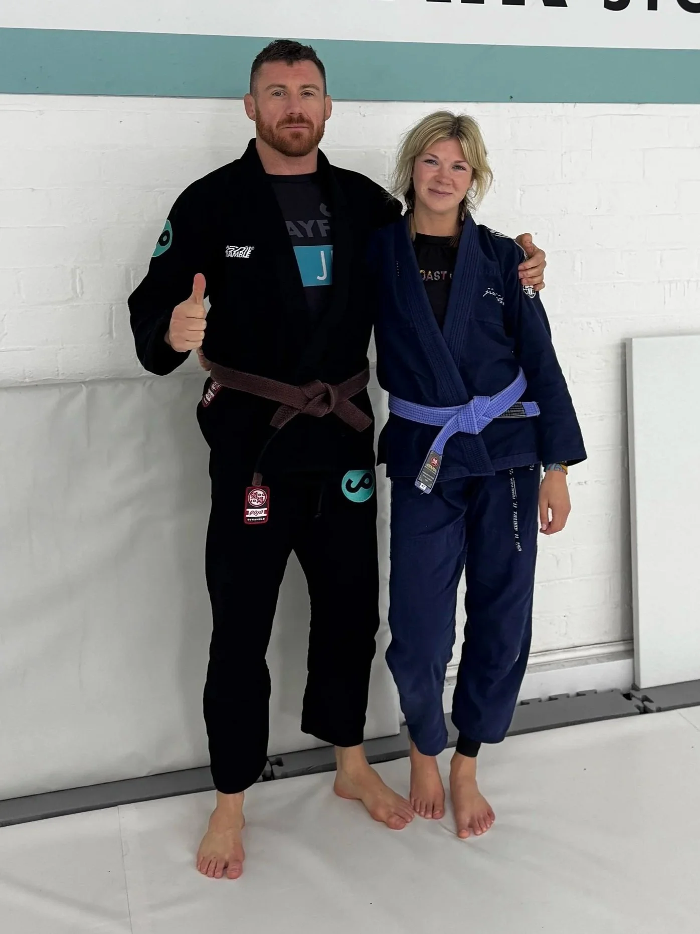 Two people in Brazilian Jiu-Jitsu Gi uniforms standing on a mat, with one giving a thumbs-up and the other smiling, in a martial arts gym. One person has a brown belt, the other a purple belt.