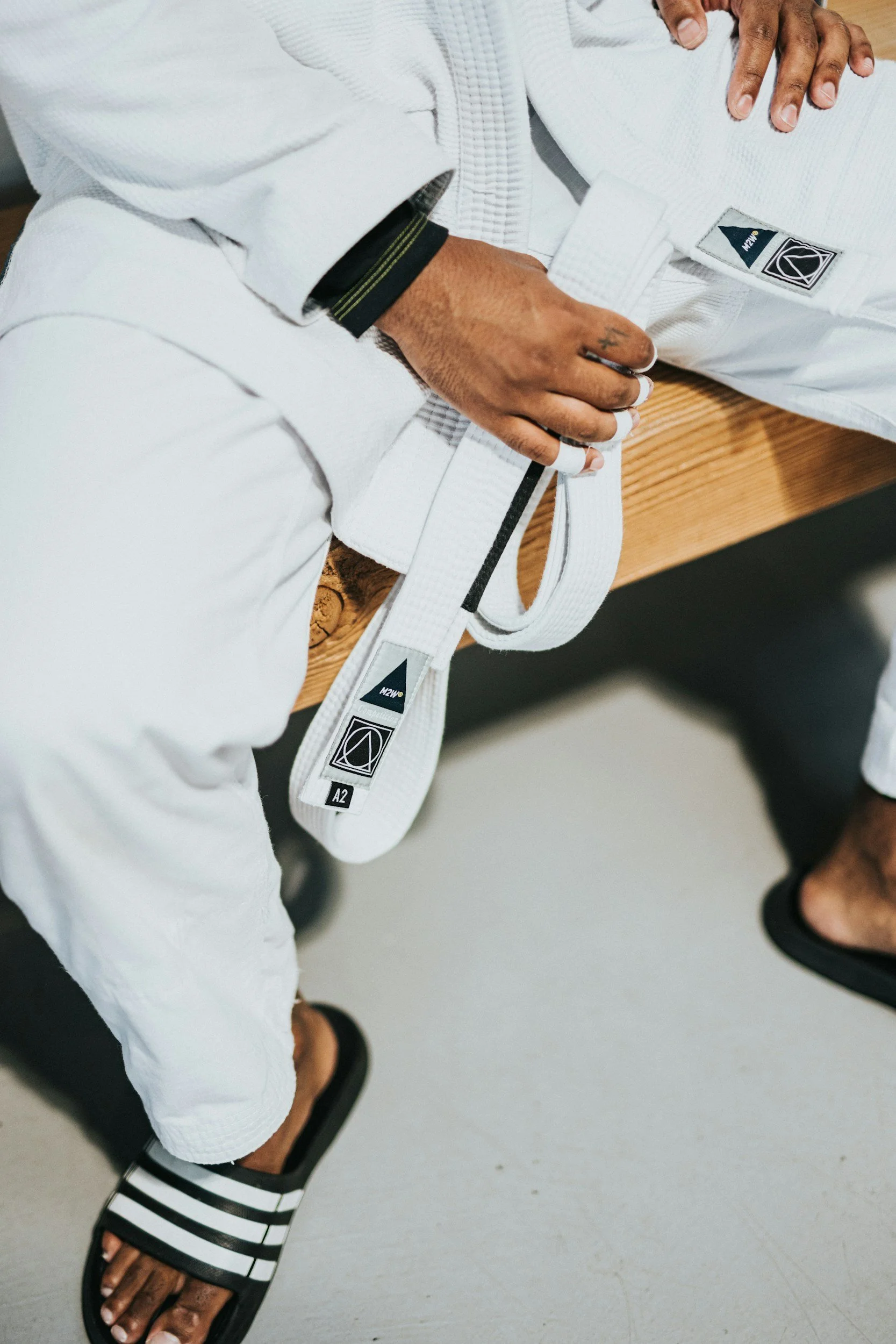A person dressed in a white martial arts gi with a white belt, sitting on a wooden bench. They are wearing black and white striped slide sandals, and their hand rests on their stomach while the other hand holds the end of the belt.