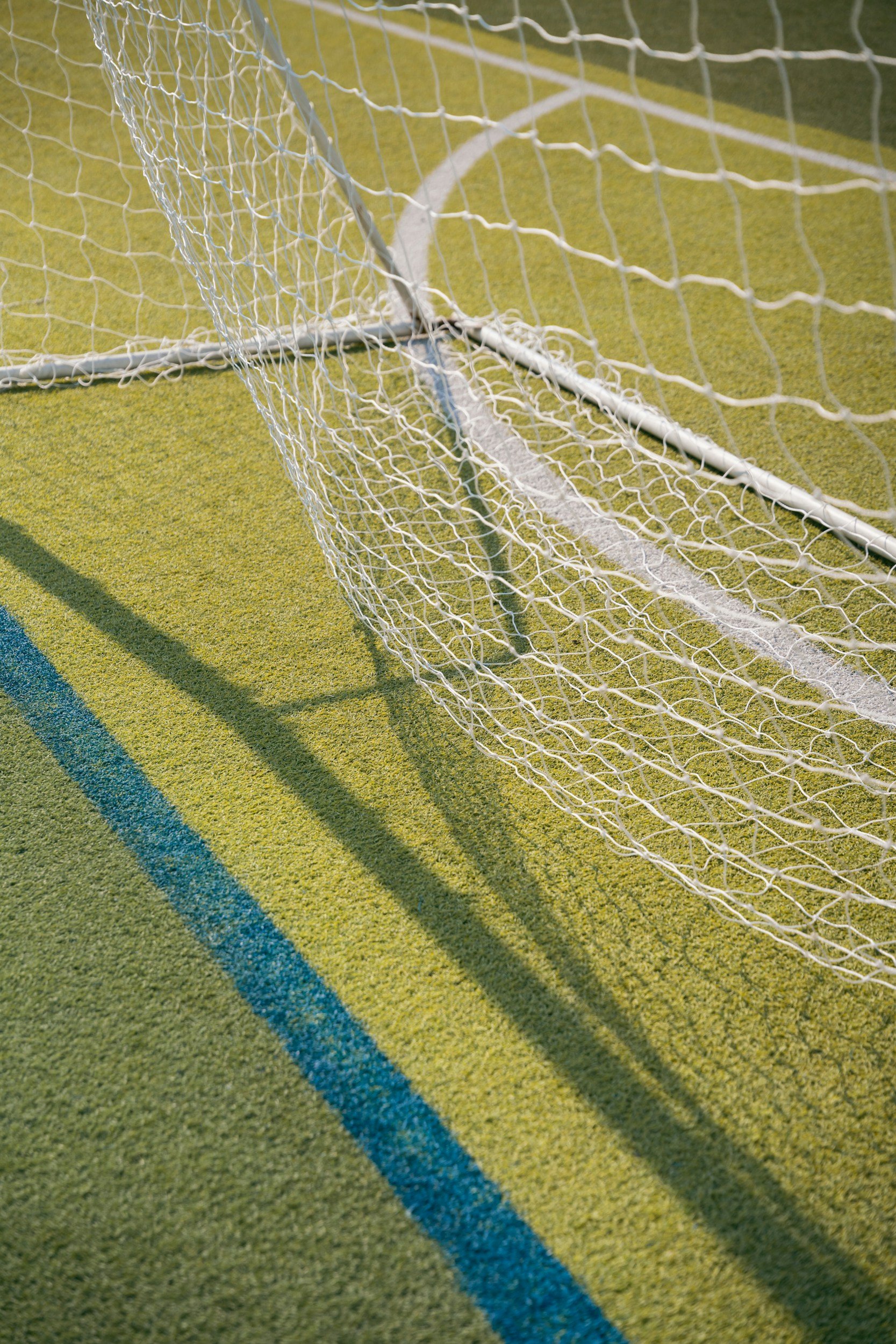 Close-up of a soccer goal net on a green artificial turf field with blue and yellow lines, casting shadows on the ground.