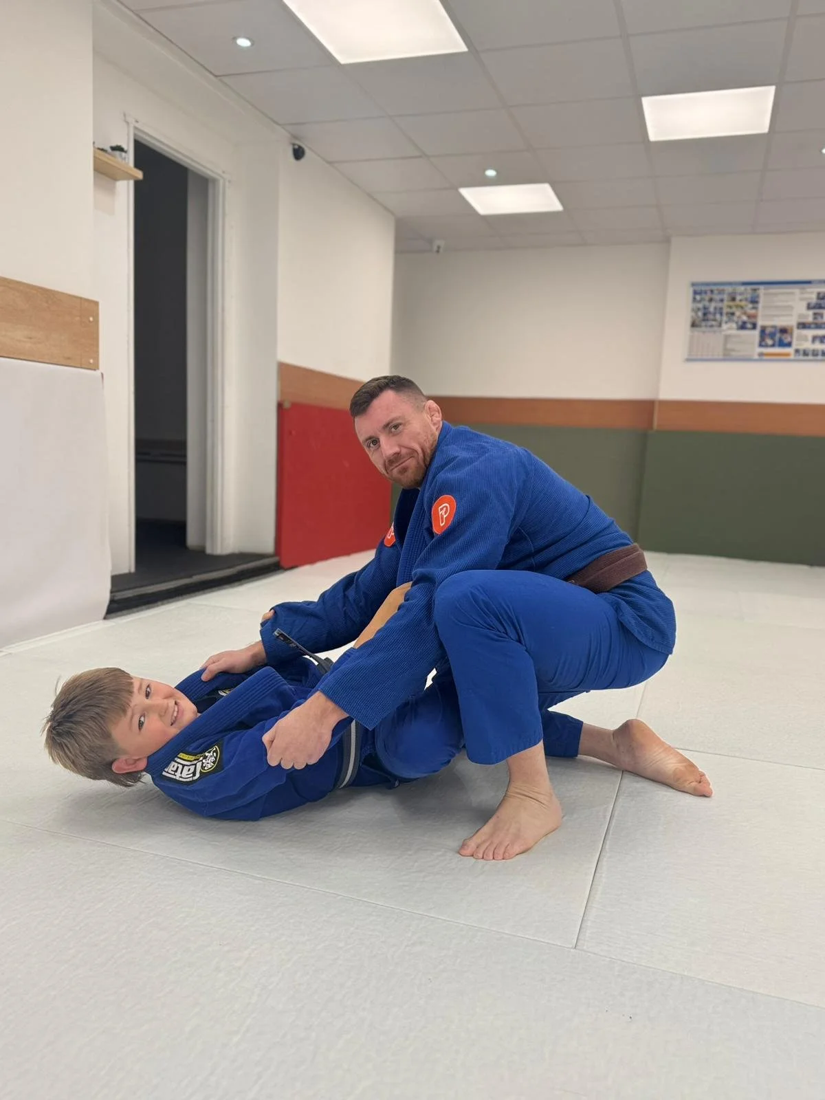 An adult man and a young boy practicing Brazilian Jiu-Jitsu on a white mat in a martial arts gym. The man is in a blue gi and is holding the boy in a grappling position, both smiling.