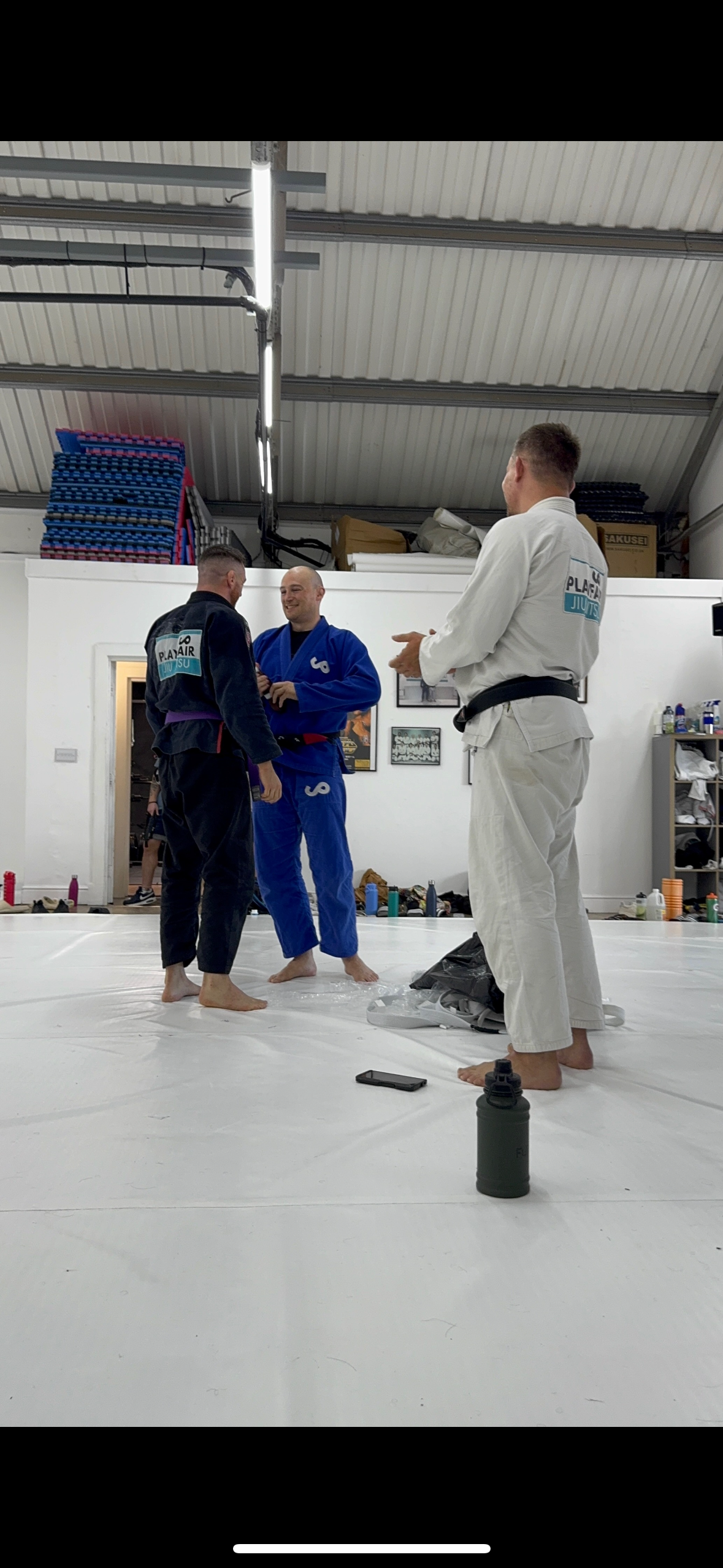 Three men in Brazilian Jiu-Jitsu gis standing and talking on a padded mat inside a gym. One is in a white gi, the other two are in black and blue gis. The gym has a white wall with framed pictures, shelves, and equipment in the background.
